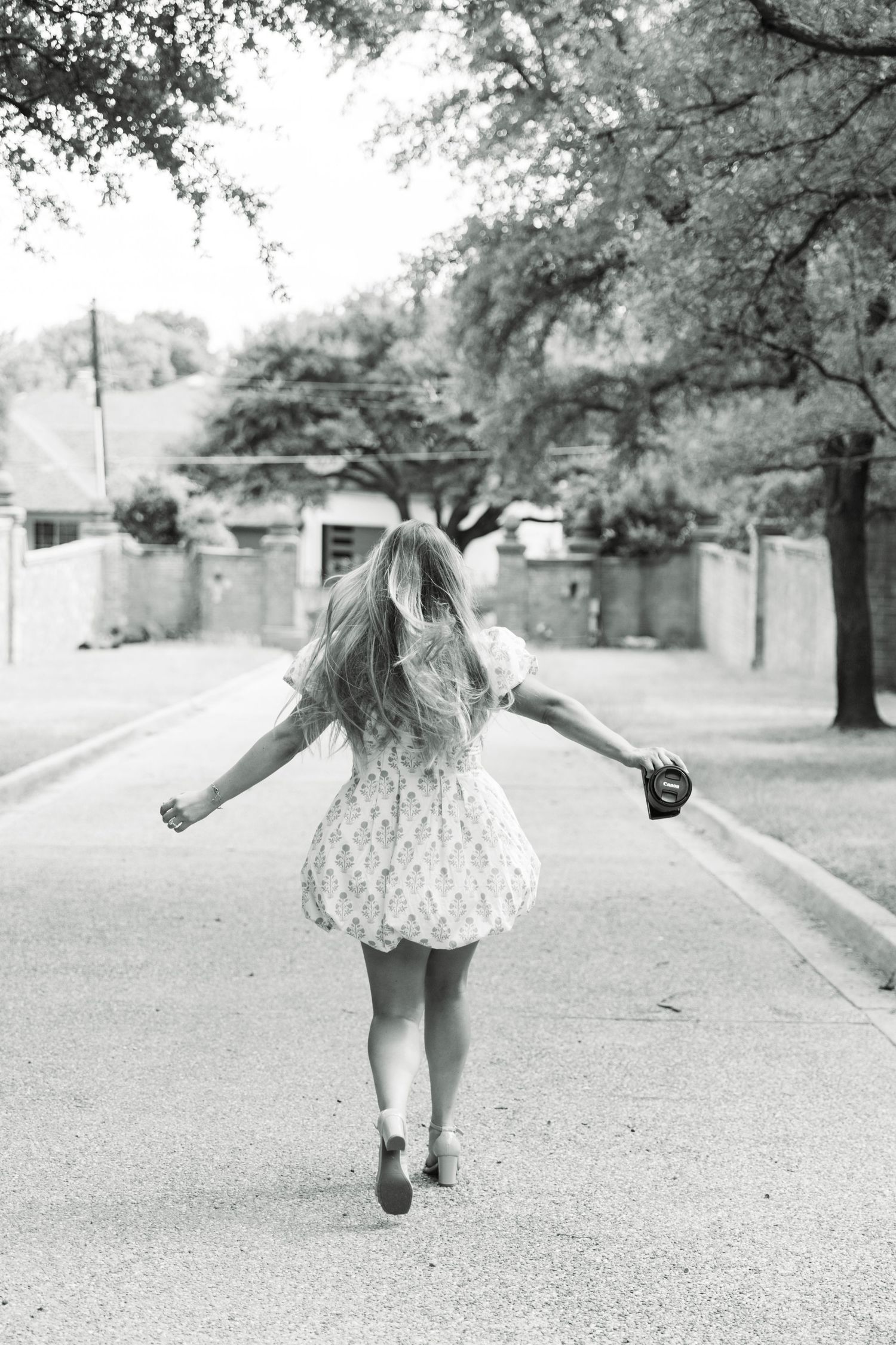 Black and white photo of a girl in a short dress walking down a tree-lined street with arms outstretched.