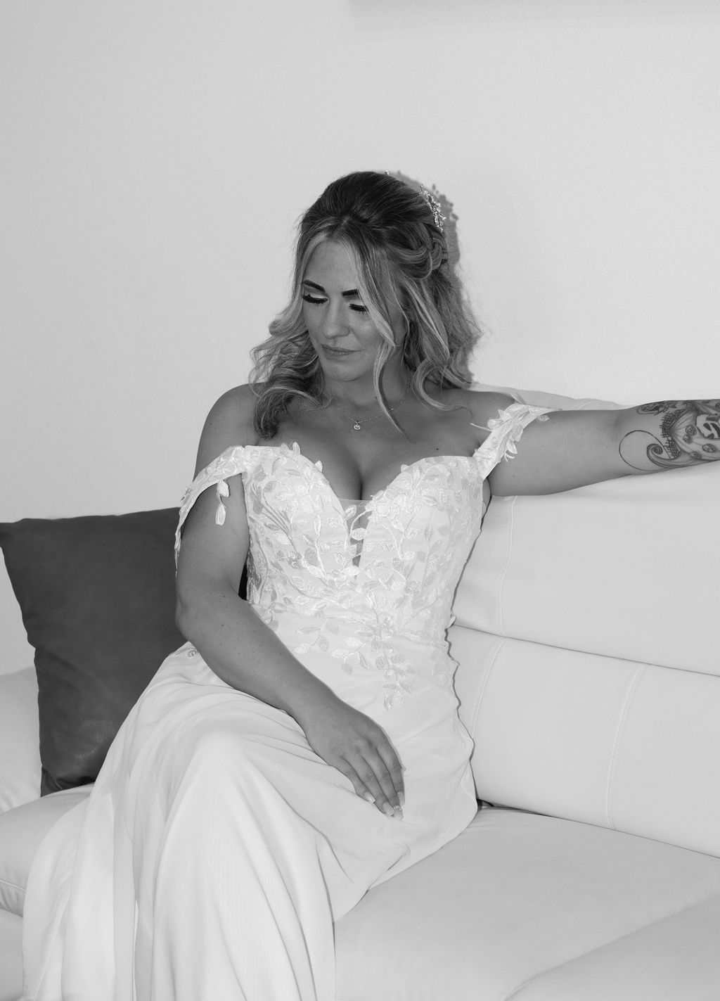 Black and white photo of a bride in an off-shoulder lace wedding gown sitting on a white sofa.