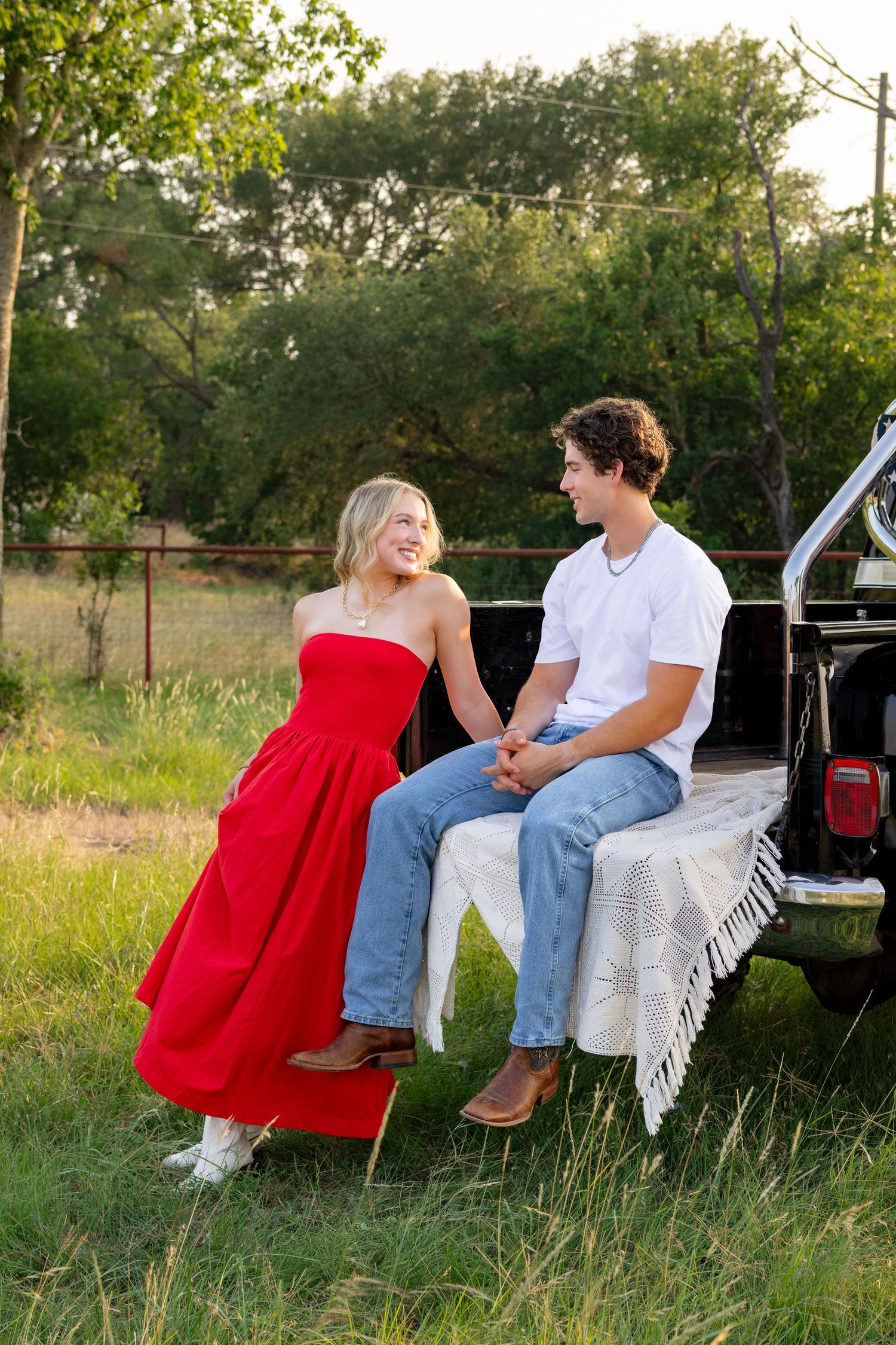A young couple holds hands on a truck tailgate, she in a red dress, he in a white tee and jeans, outdoors.
