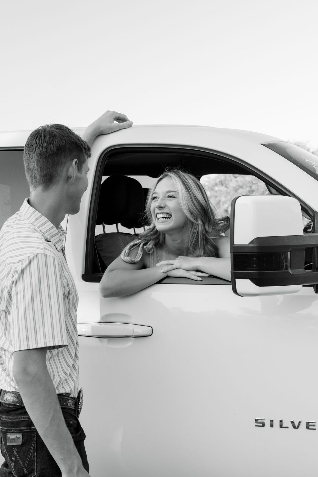Black and white photo of a laughing woman leaning out of a Chevy Silverado truck window talking to a young man.