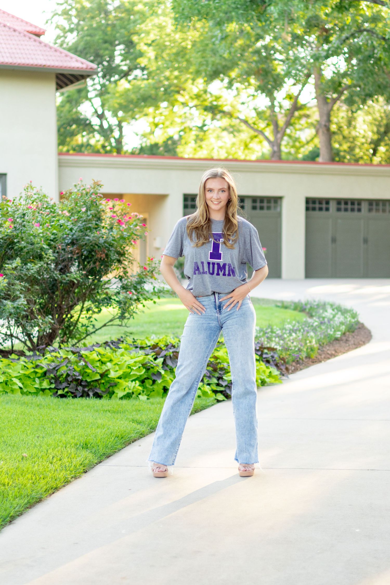 Young woman in gray alumni t-shirt and jeans posing on driveway in front of suburban home with lush landscaping.