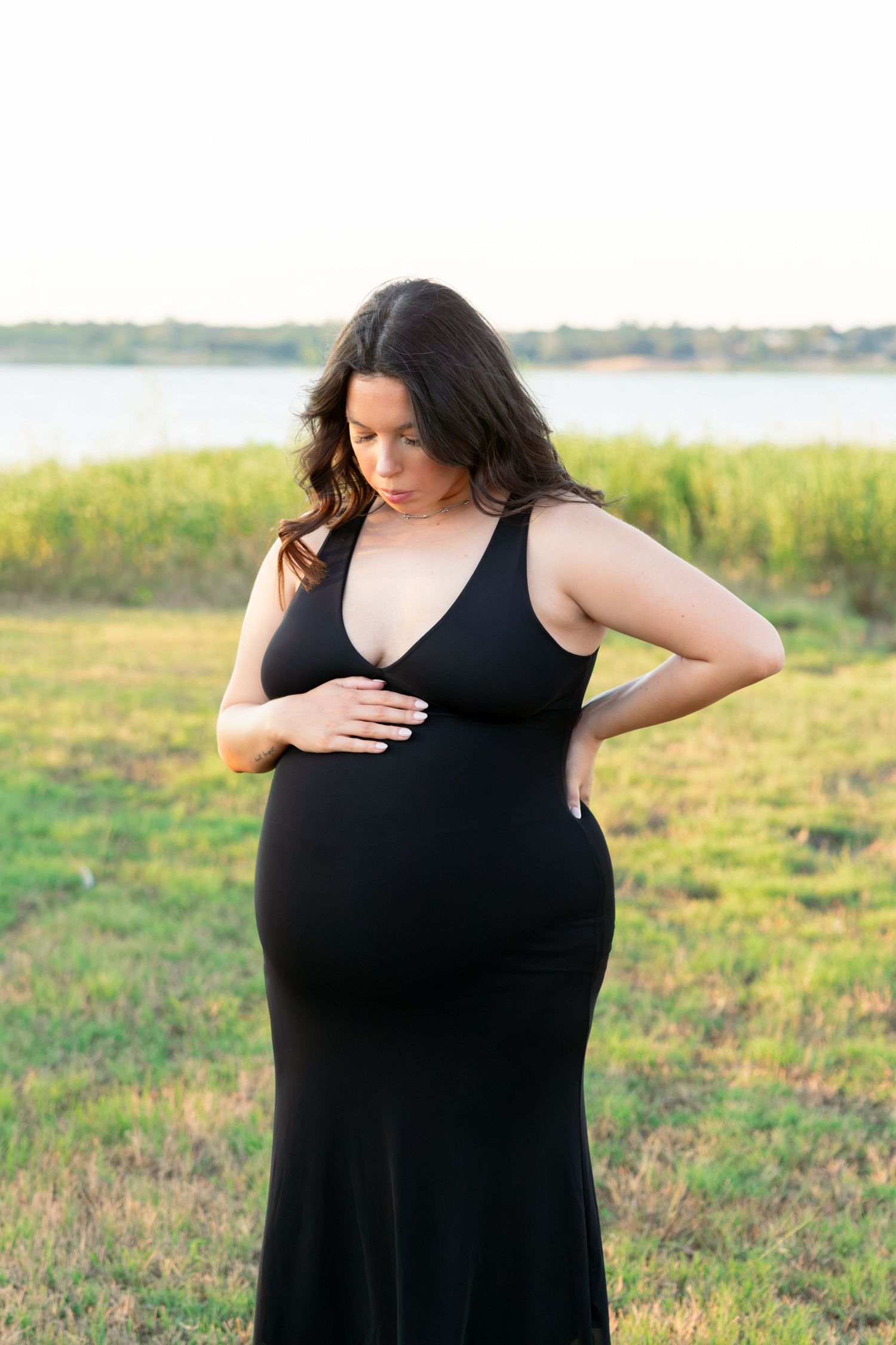Pregnant woman in black maxi dress cradling baby bump outdoors near lake at golden hour.
