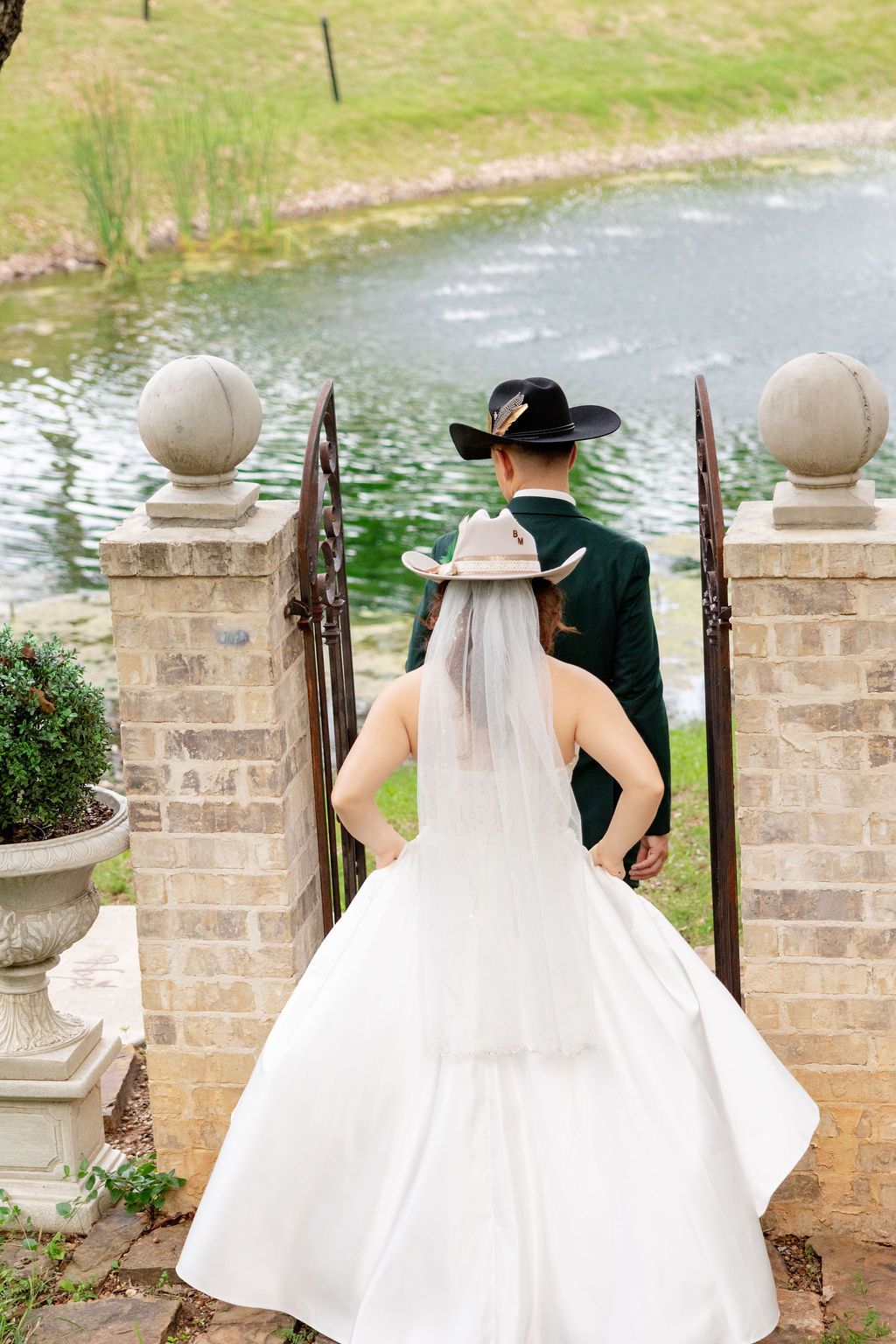 Bride and groom in cowboy hats stand at a garden gate overlooking a pond on their wedding day.