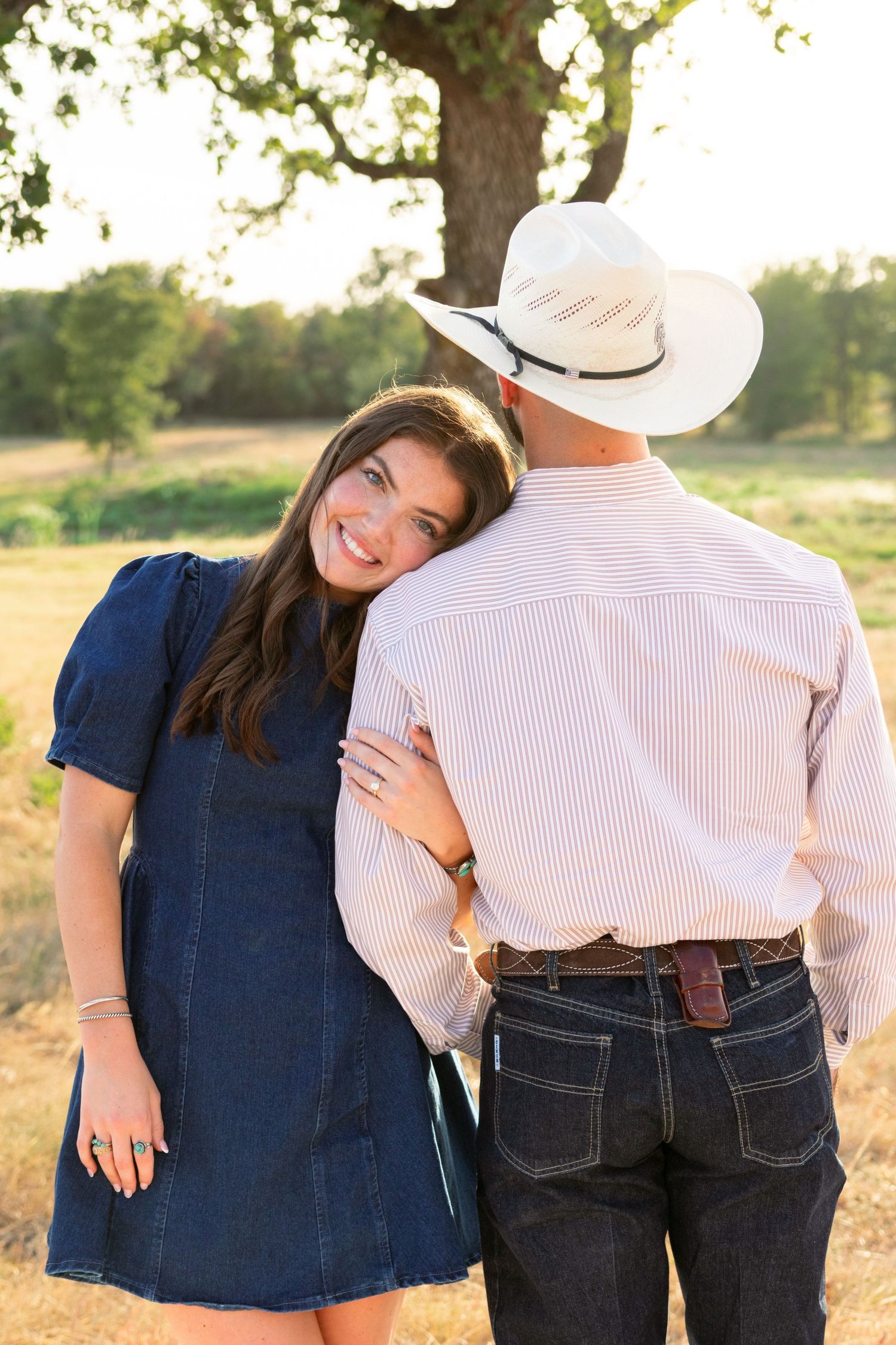 Woman in denim dress leans on man in cowboy hat outdoors.
