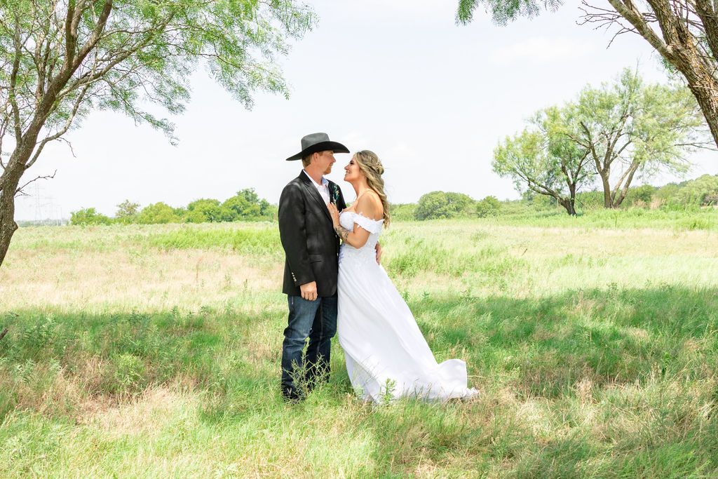 A bride and groom share a romantic moment in a lush green field, with the groom wearing a black cowboy hat and jeans.