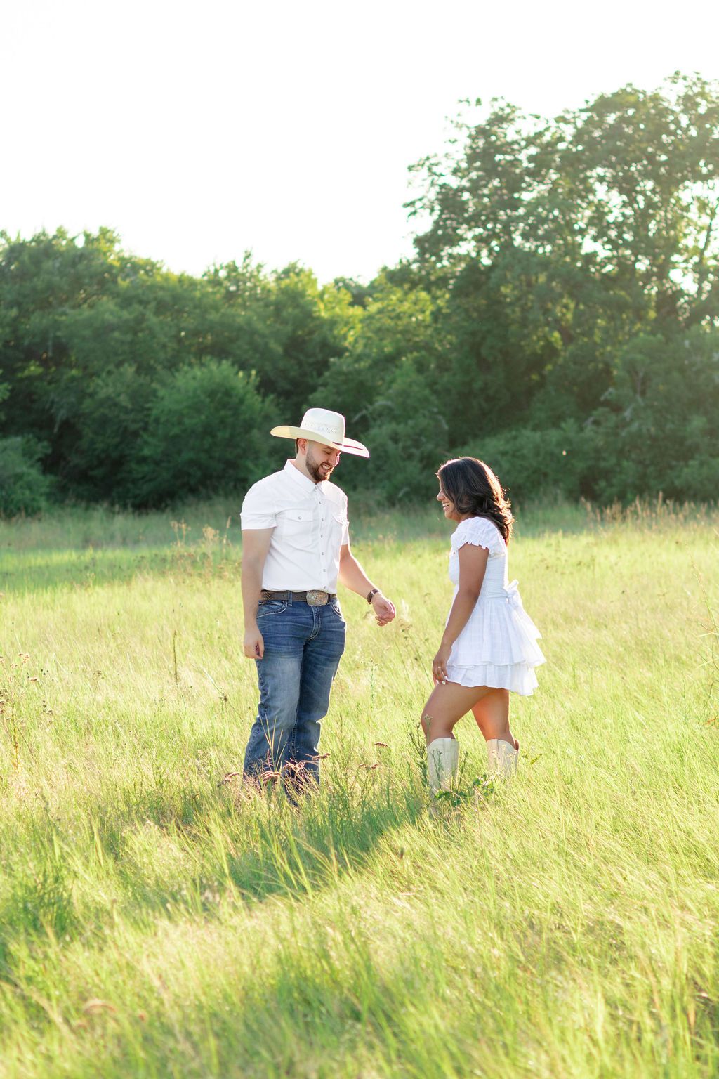 Couple holding hands in a sunlit green field, man in cowboy hat and jeans, woman in white dress and boots.
