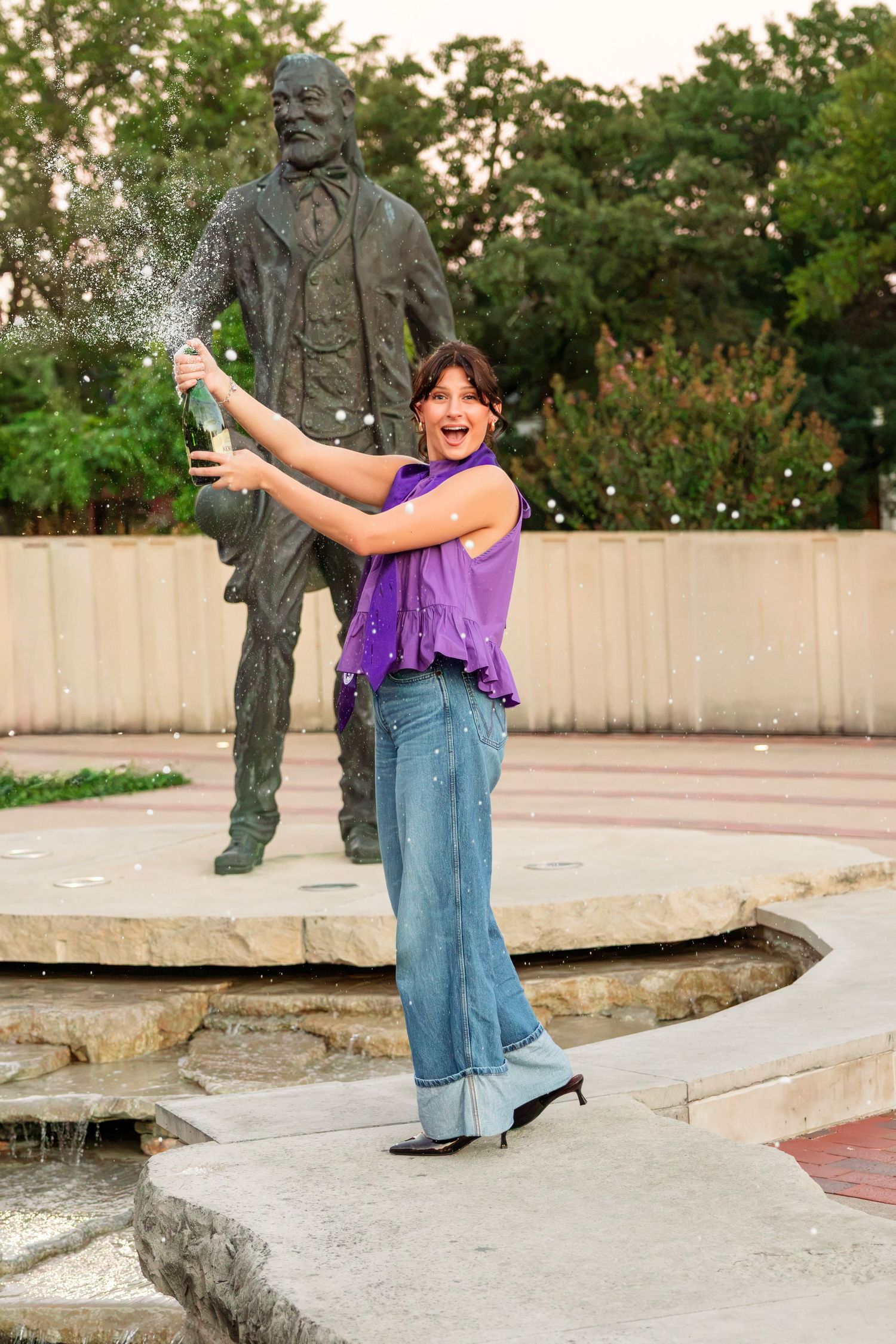 Woman in a purple top and wide-leg jeans pops a champagne bottle beside a bronze statue near a stone fountain.