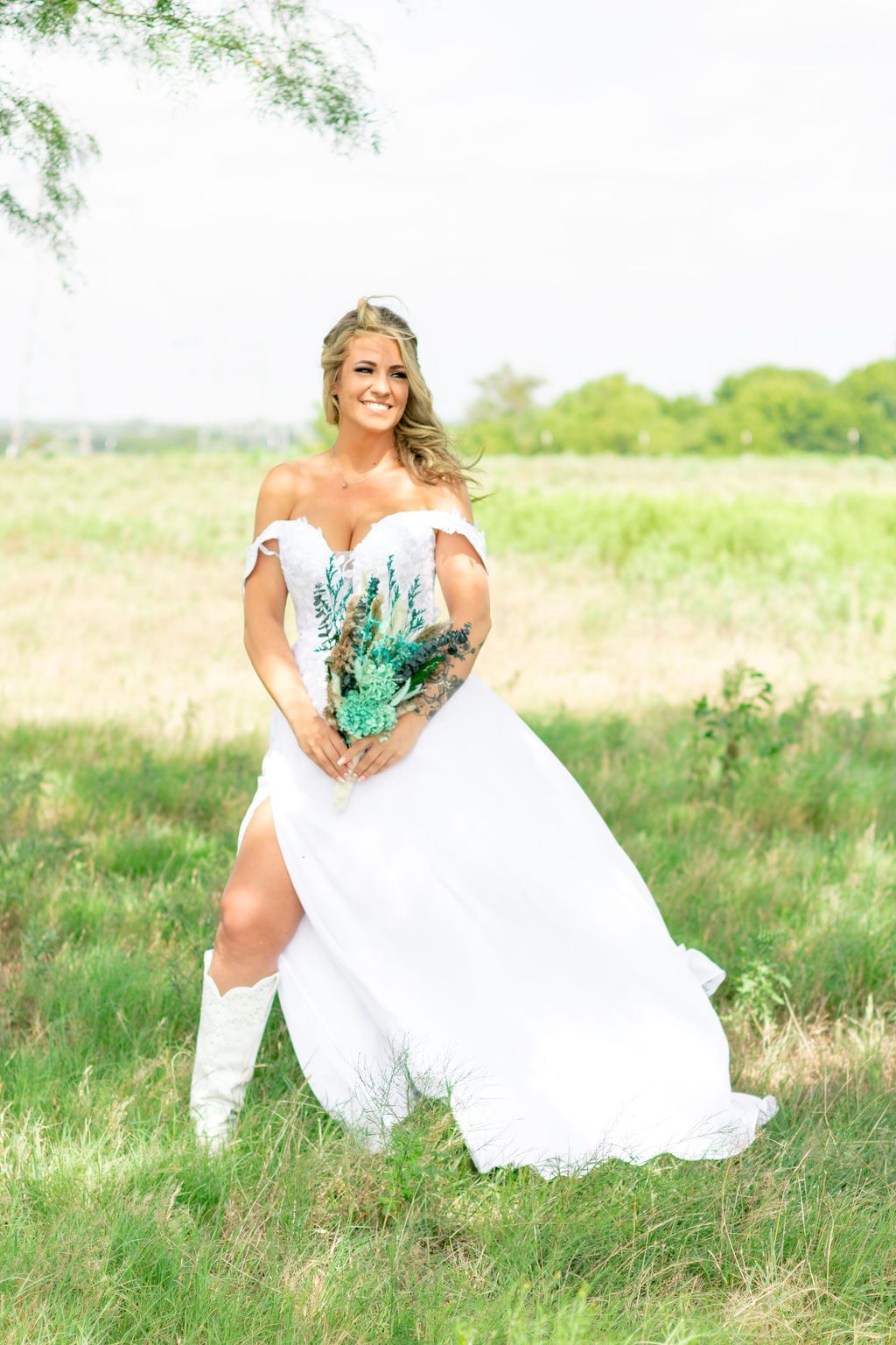 Smiling bride in off-shoulder white gown with slit, cowboy boots, holding teal bouquet in a green field.