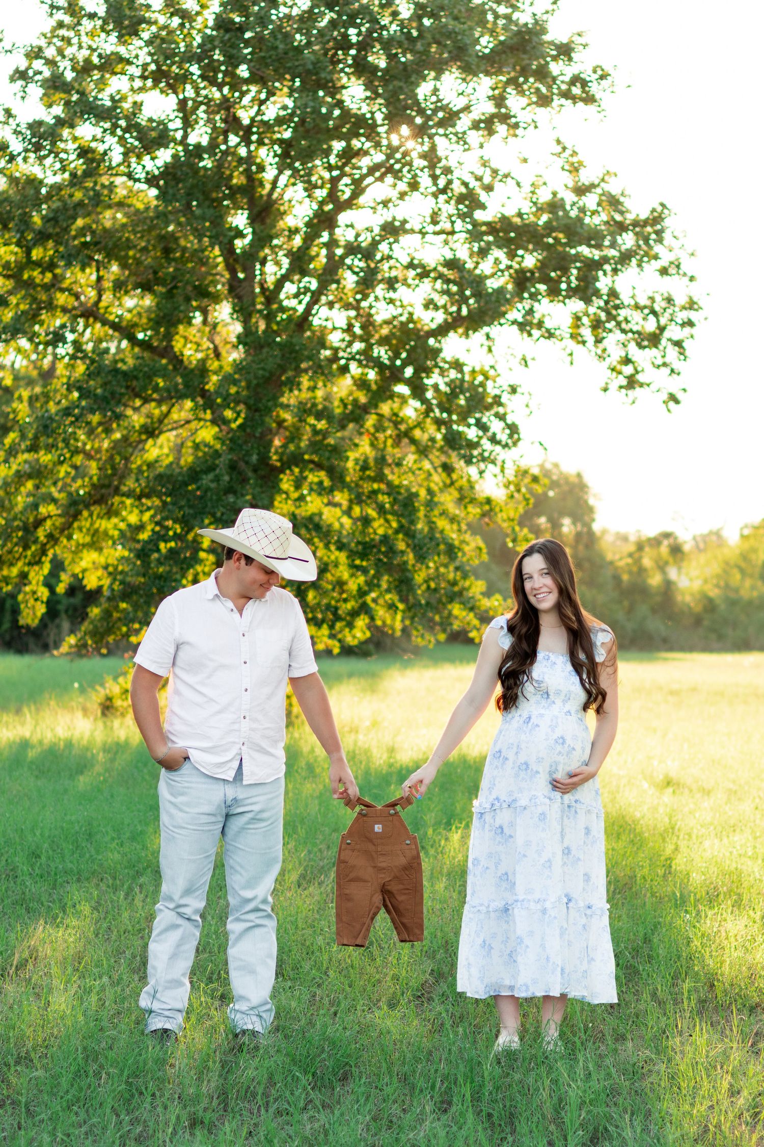 Expecting couple in a sunny grassy field holding a tiny brown baby overall between them, announcing their pregnancy beneath a large tree.