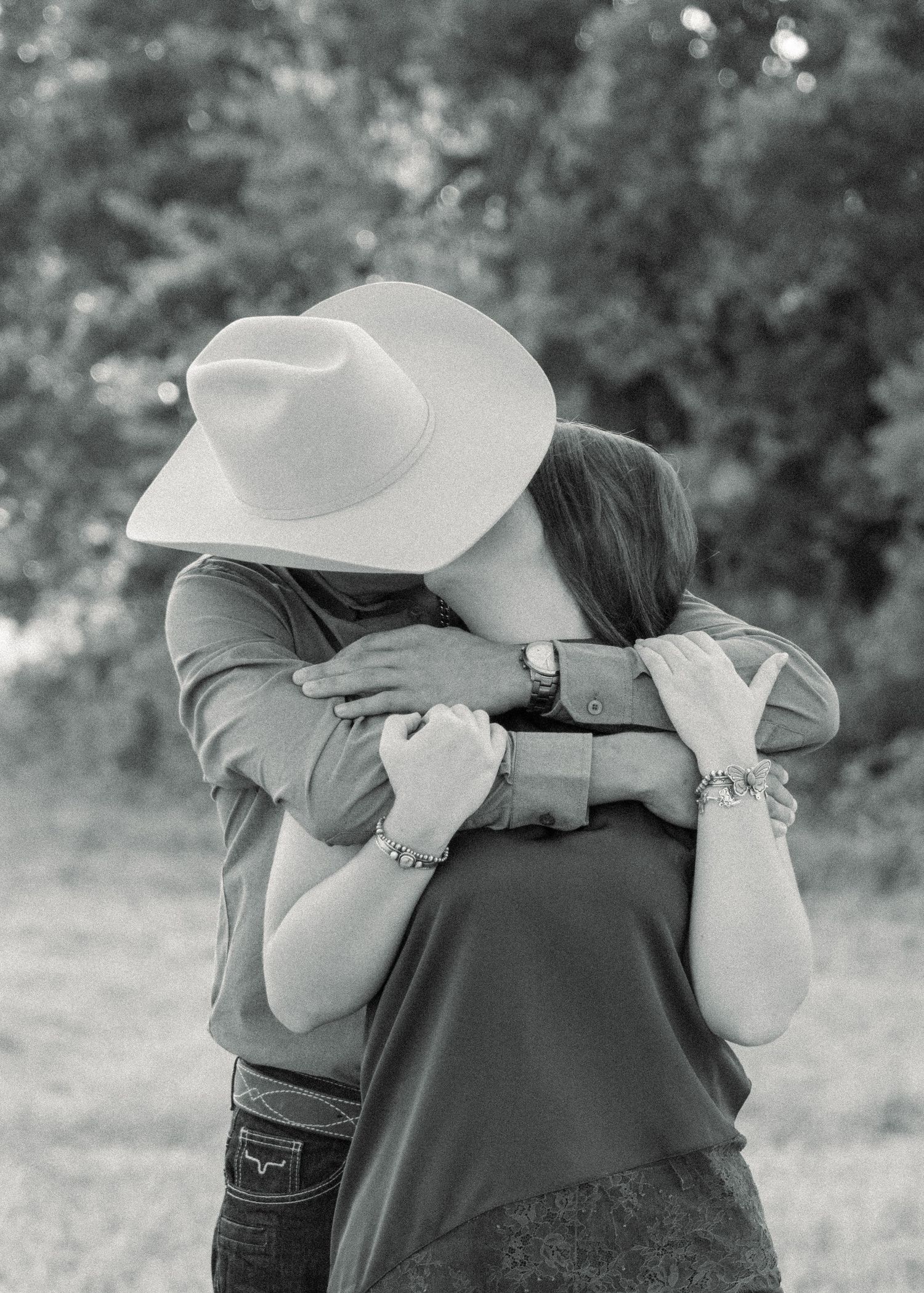 Black and white photo of a couple embracing outdoors, man wearing a cowboy hat, woman in a casual top with bracelets.