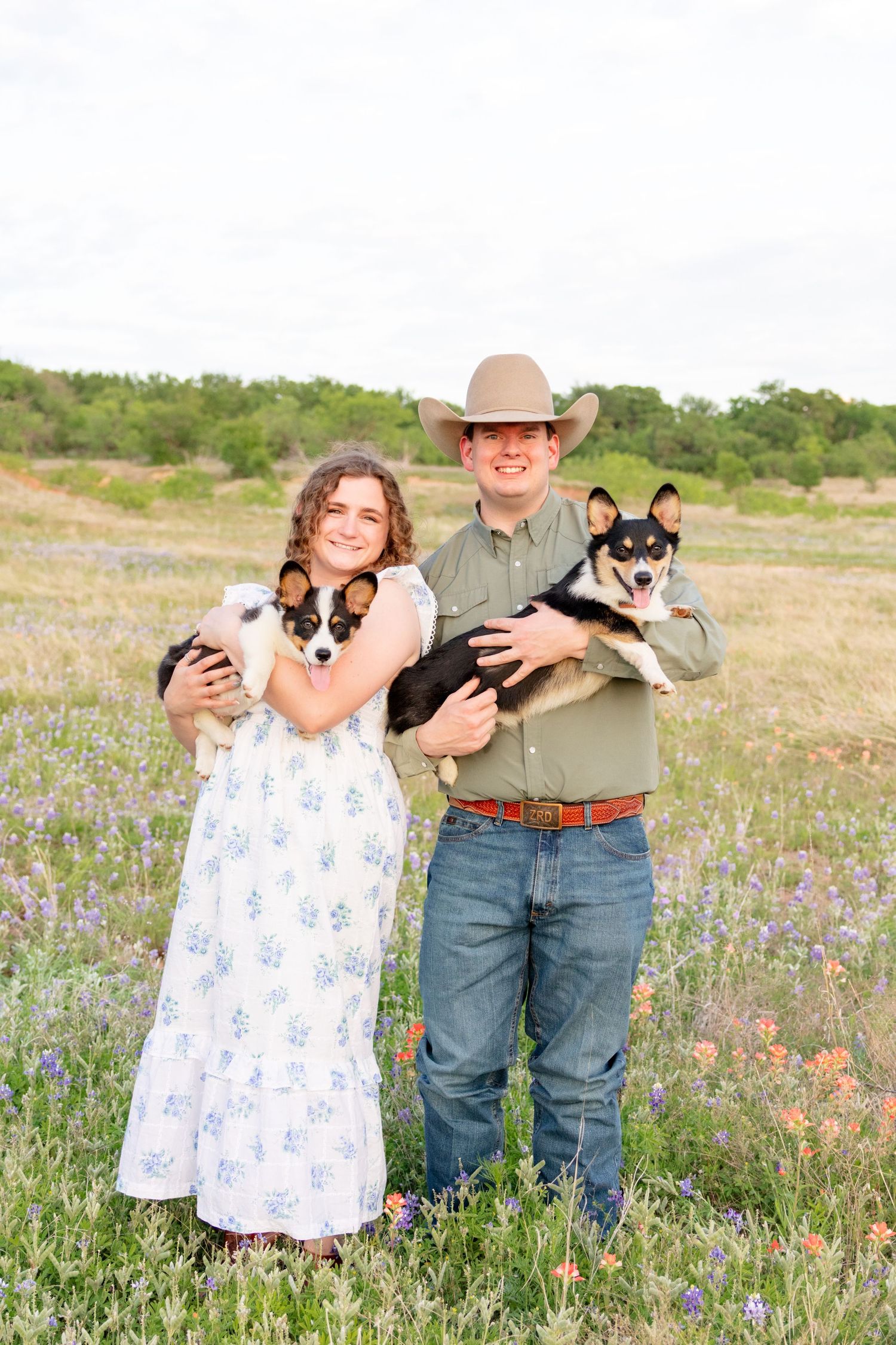 Two smiling kids holding Corgi dogs in a wildflower field, boy wearing cowboy hat and girl in floral dress.  Granbury Wedding Photographer