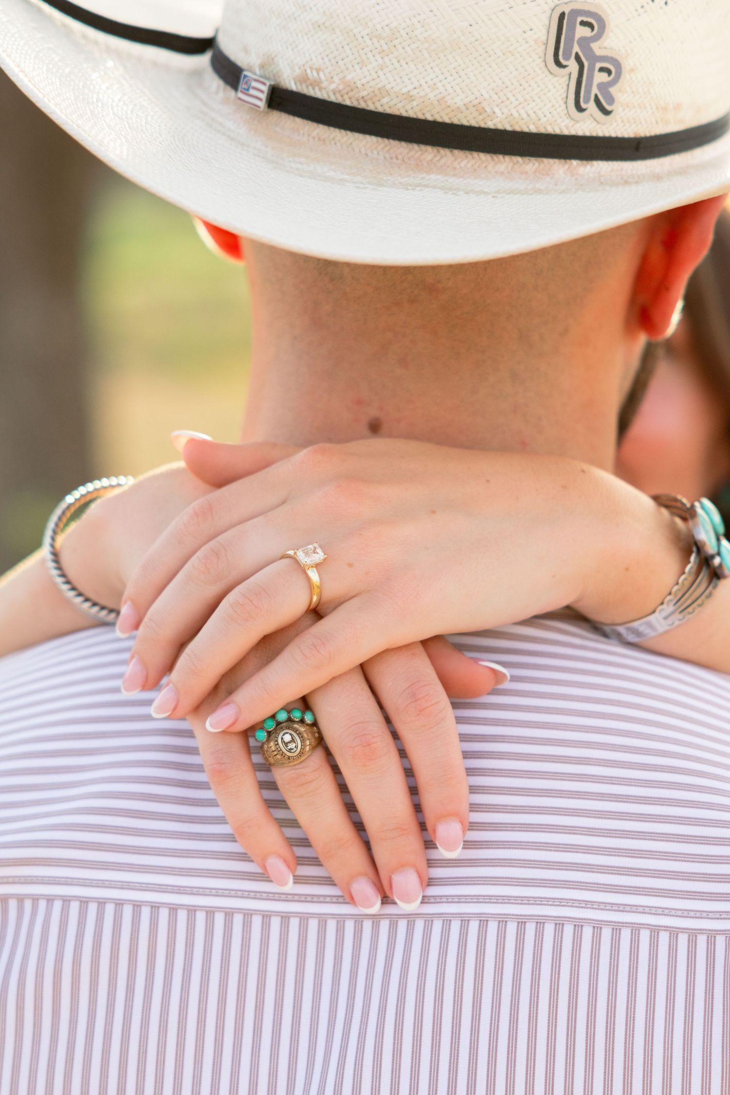Close-up of woman's hands with diamond engagement ring and turquoise ring resting on man's back in cowboy hat.