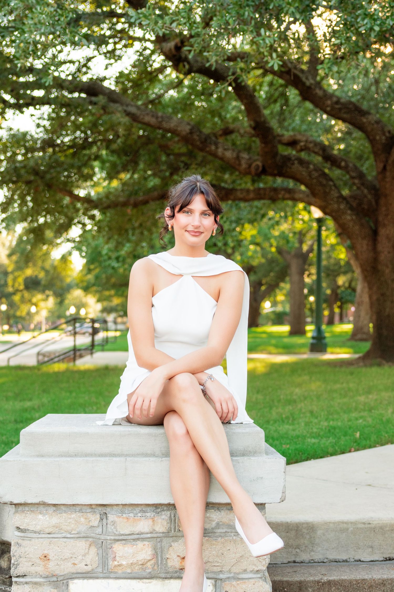 Woman in a white halter dress and white heels sits with legs crossed on a stone ledge beneath large oak trees in a sunny park.
