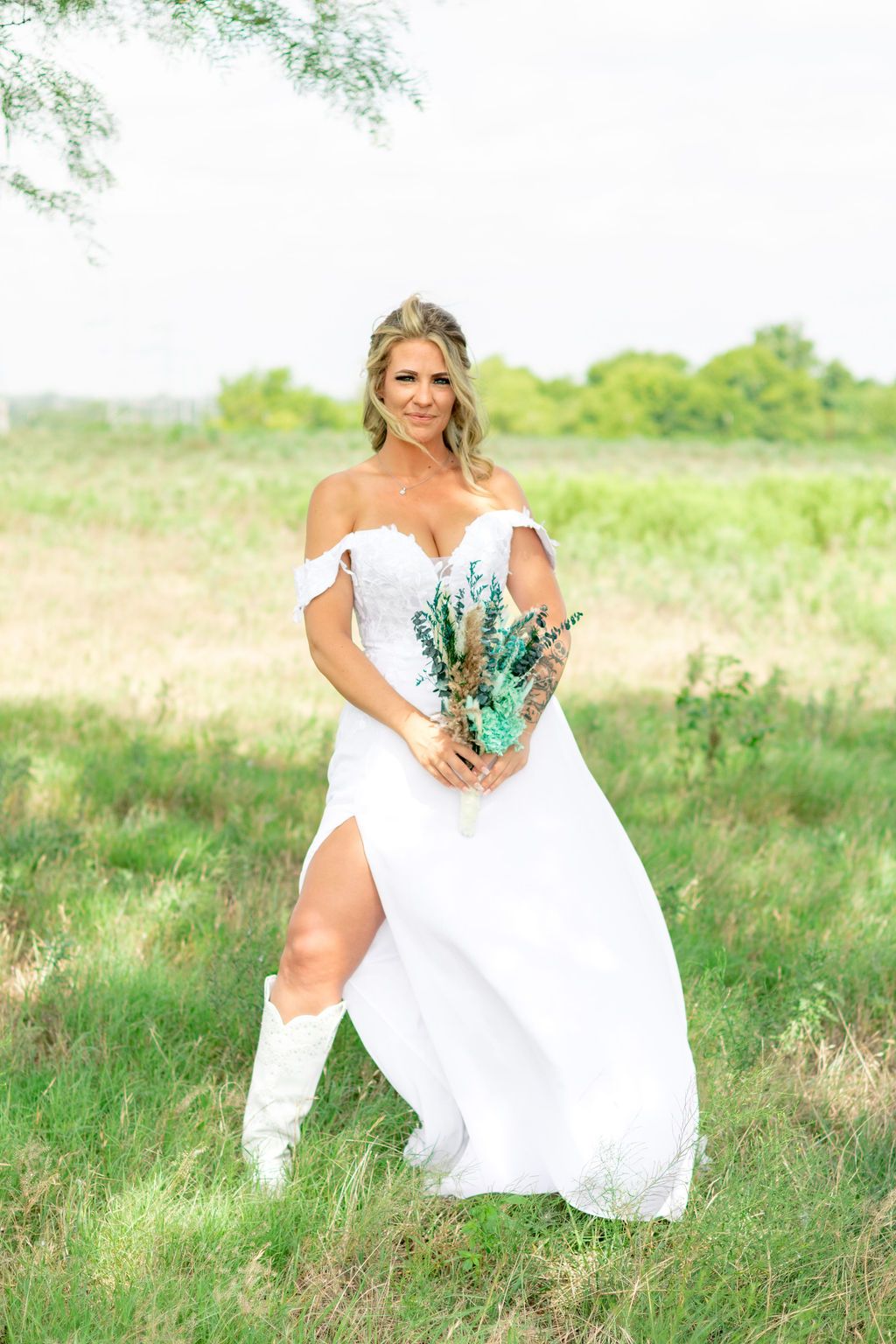 Bride in white off-shoulder gown and cowboy boots holding teal bouquet in green field.