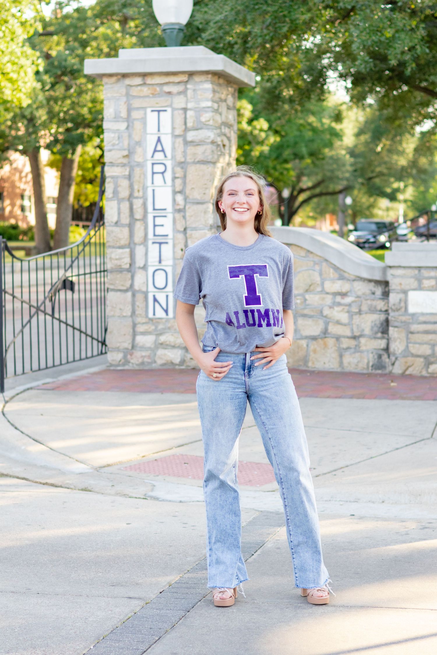 Smiling Tarleton State University alumni woman in gray t-shirt and jeans posing at campus entrance gate.