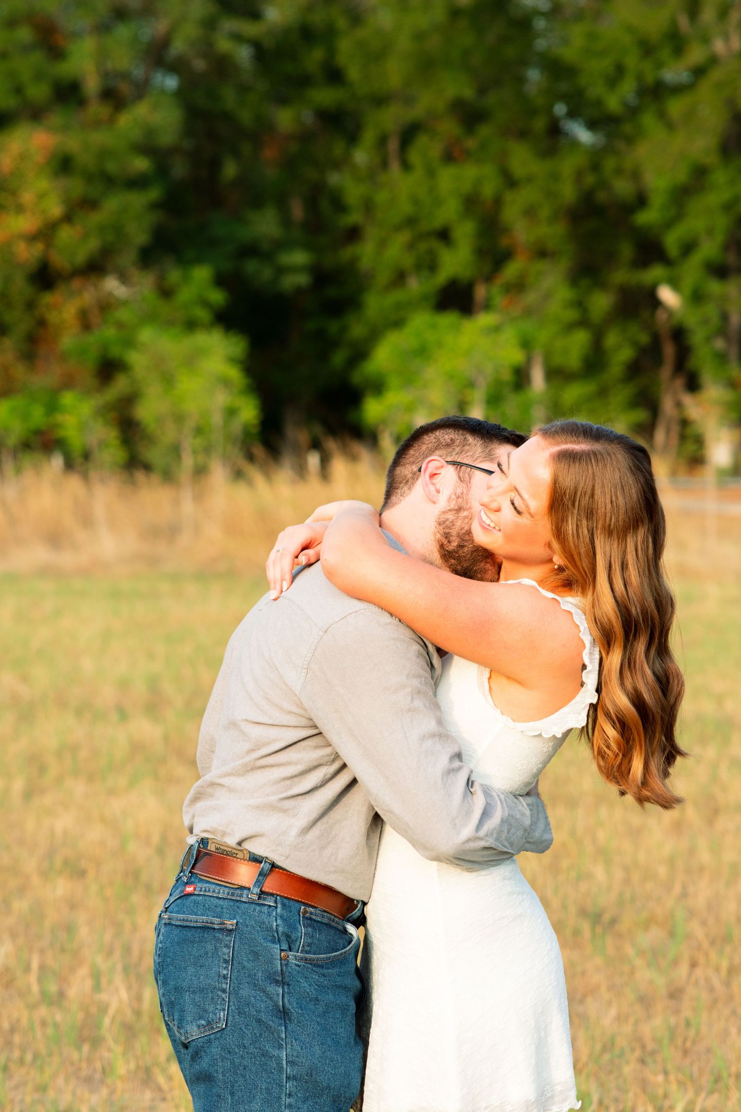 Couple embracing and kissing in a sunlit field with green trees, woman in white dress, man in jeans and gray shirt.