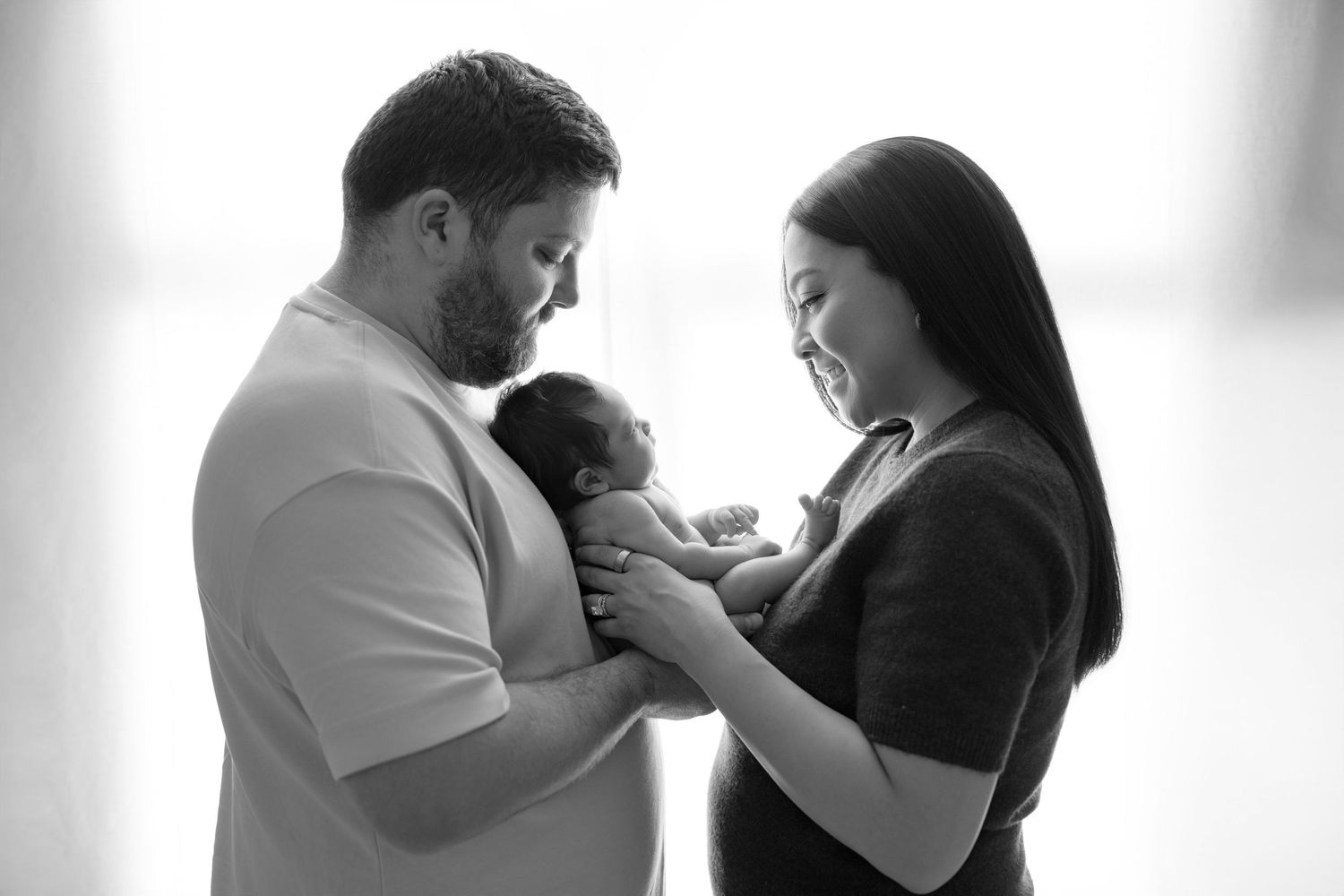 Black and white photo of new parents lovingly cradling their newborn baby together in a bright, serene setting.