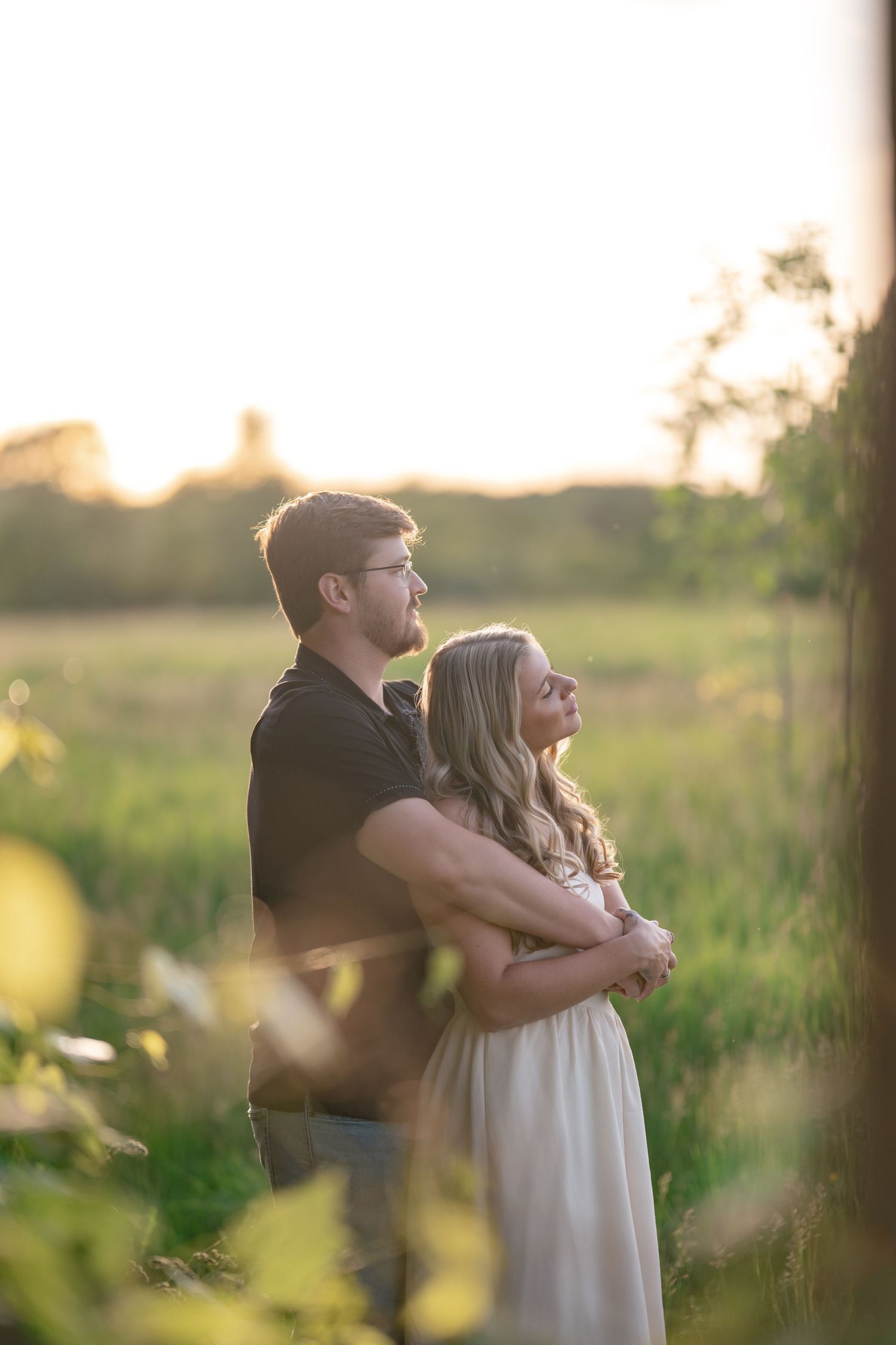 Couple embracing at golden hour in a green field, man hugging woman from behind as they gaze into the sunset.