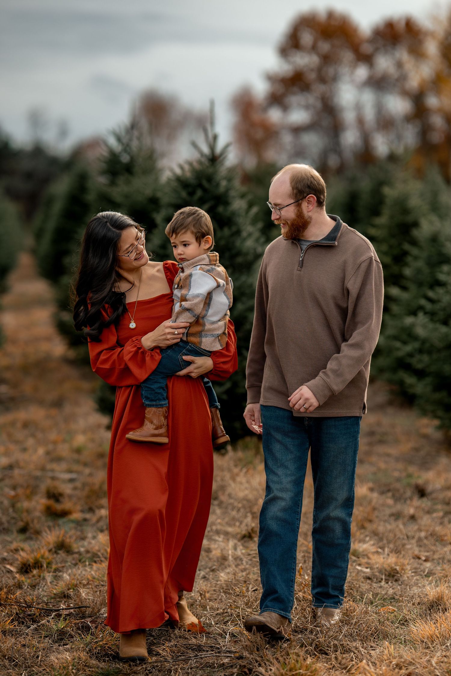Family of three posing at a Christmas tree farm, mother in red dress holding toddler, father in brown sweater and jeans.