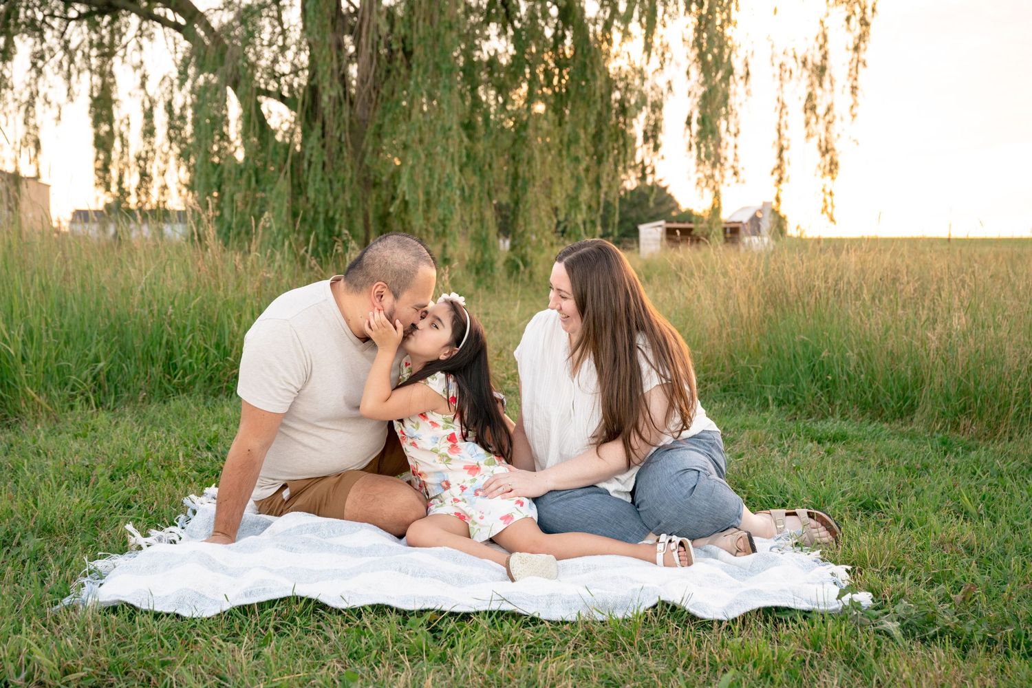 Family of three sitting on blanket in green field, parents kissing child, golden hour sunset photography.