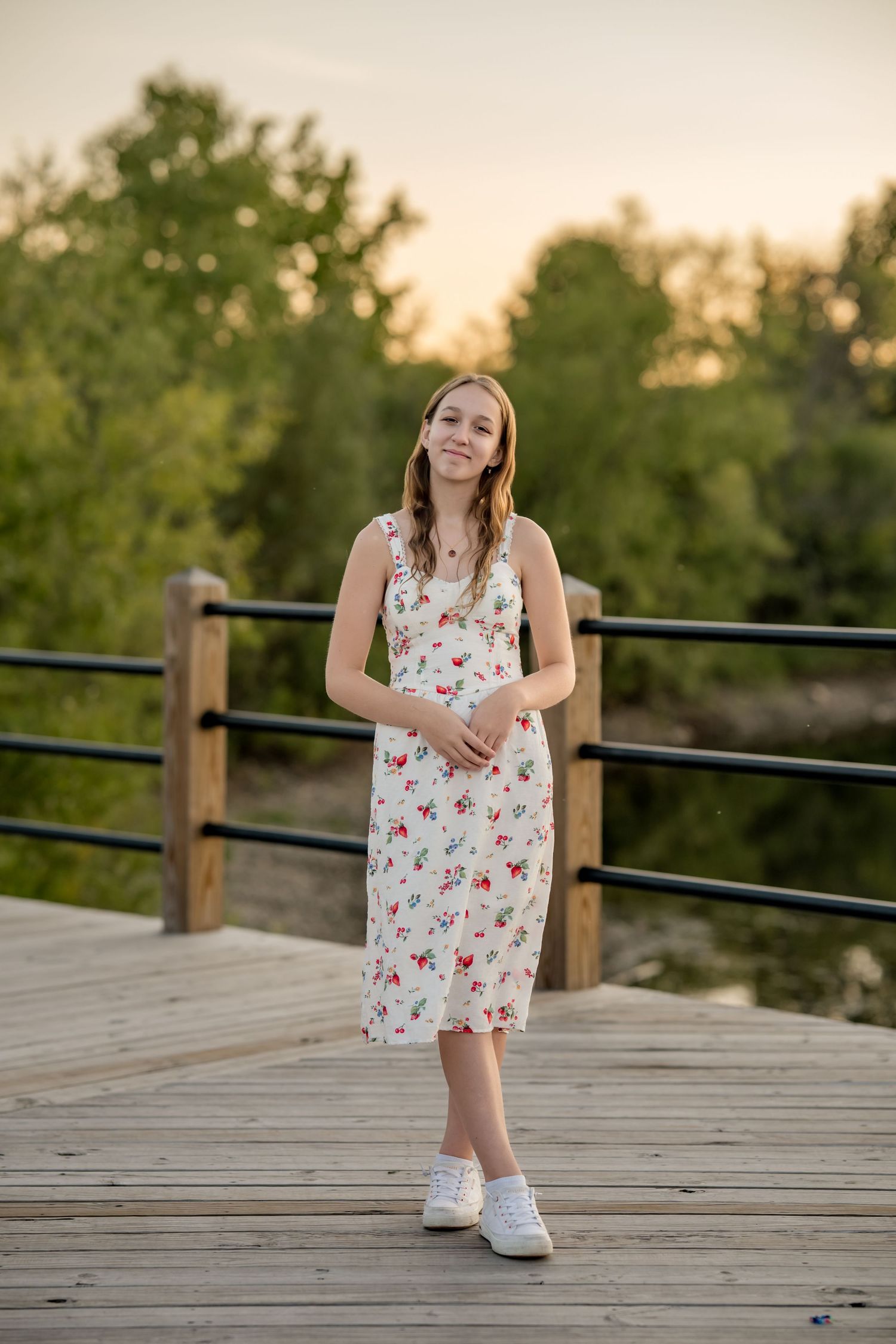 Young woman in floral midi dress standing on wooden bridge at sunset with green trees in background.