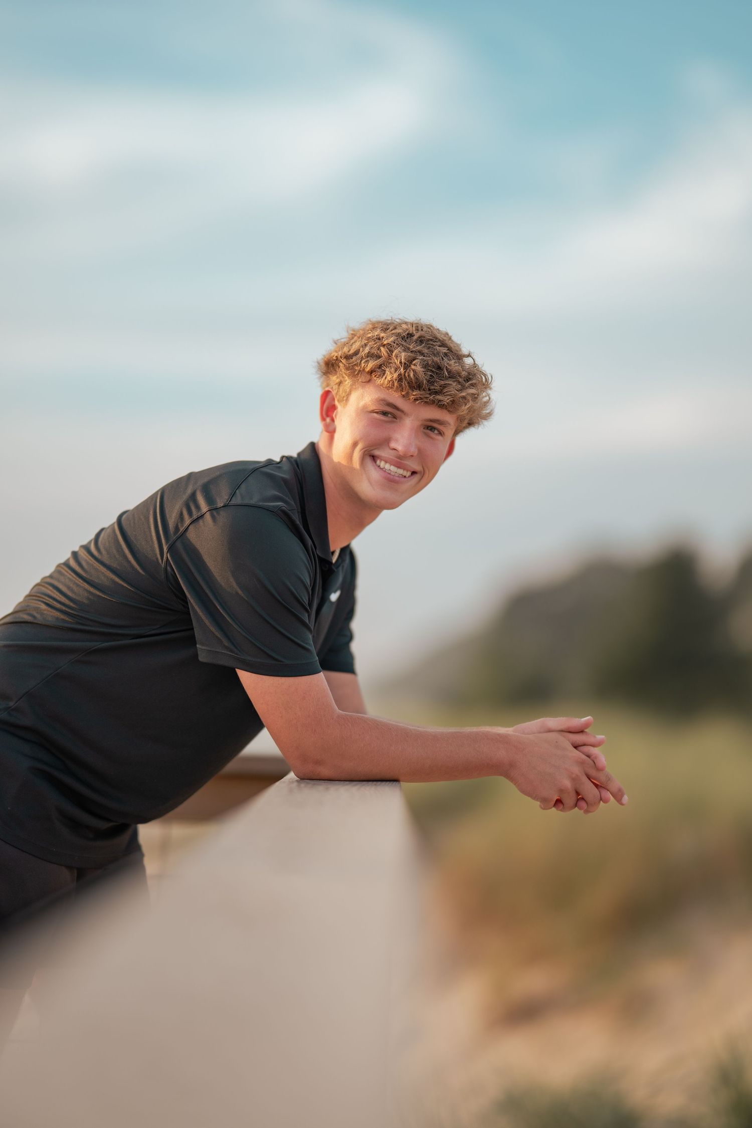 Smiling young man with curly hair leaning on a railing outdoors in a black polo shirt.
