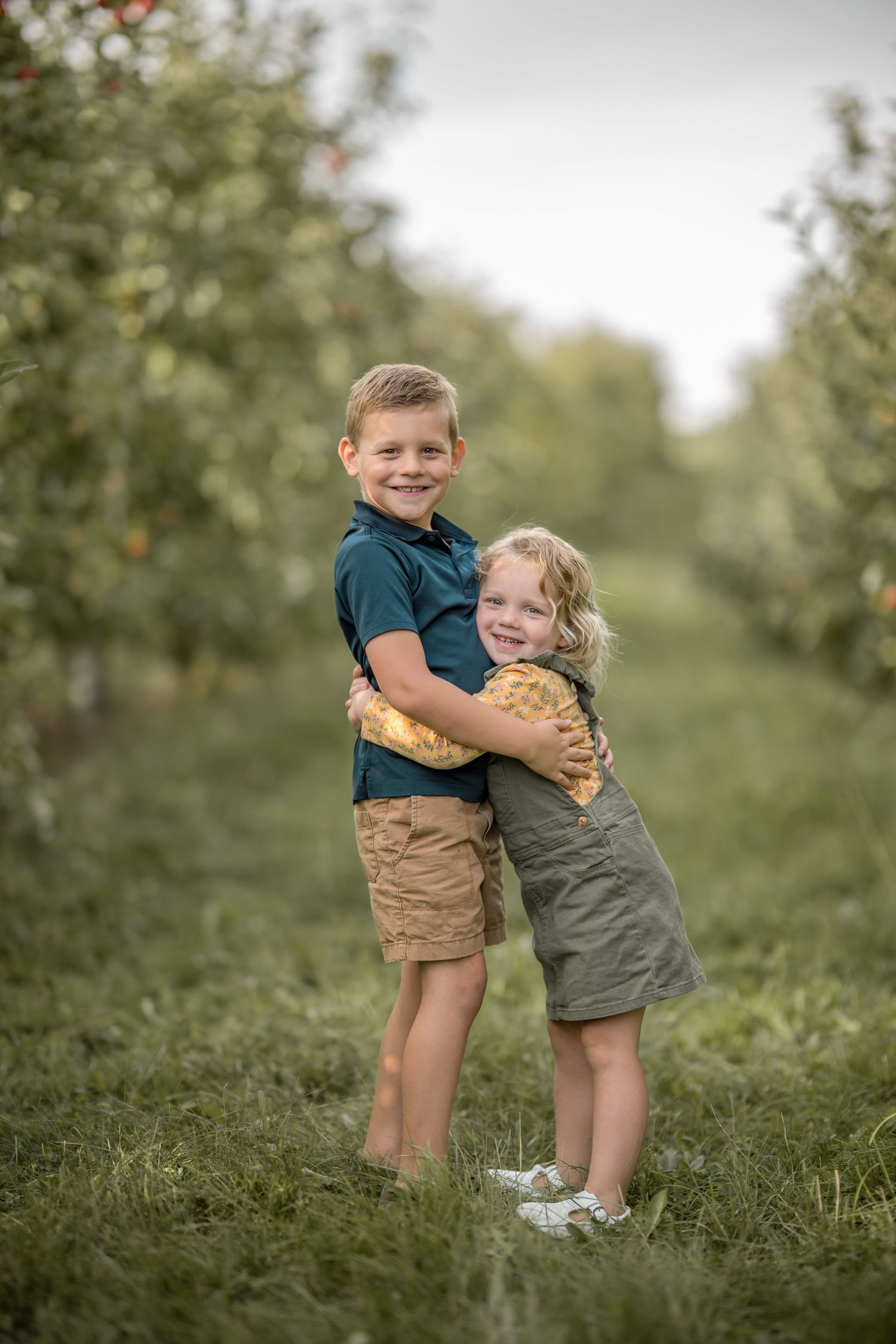 Two young siblings hugging and smiling in a lush green apple orchard during a family photo session.