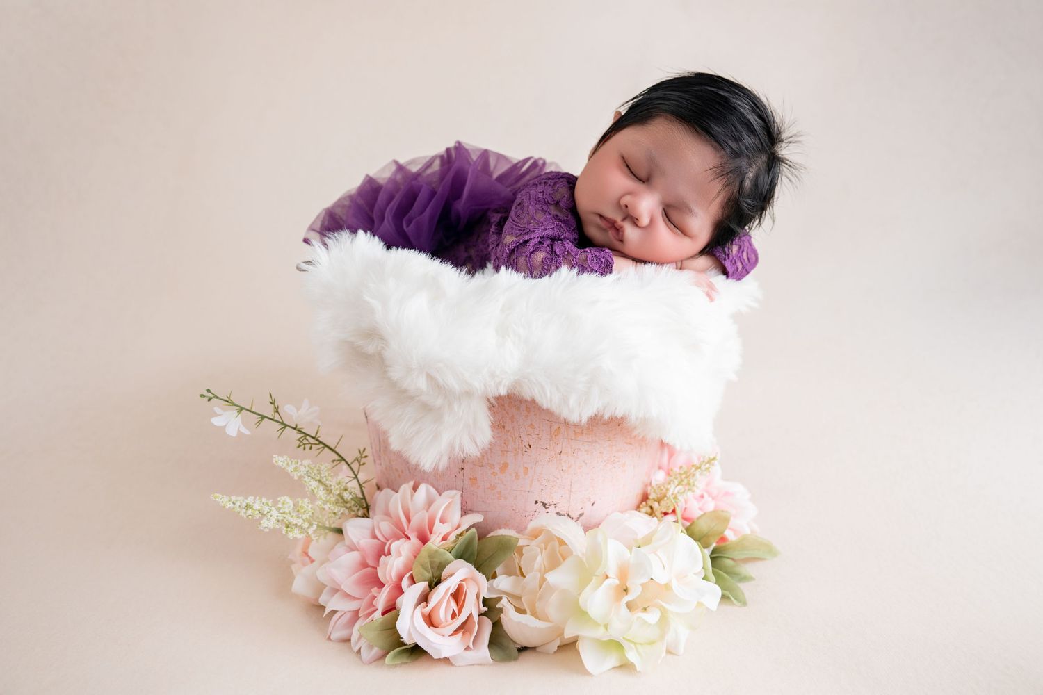 Newborn baby sleeping peacefully in a pink bucket with white feathers, wrapped in purple, surrounded by pastel flowers.