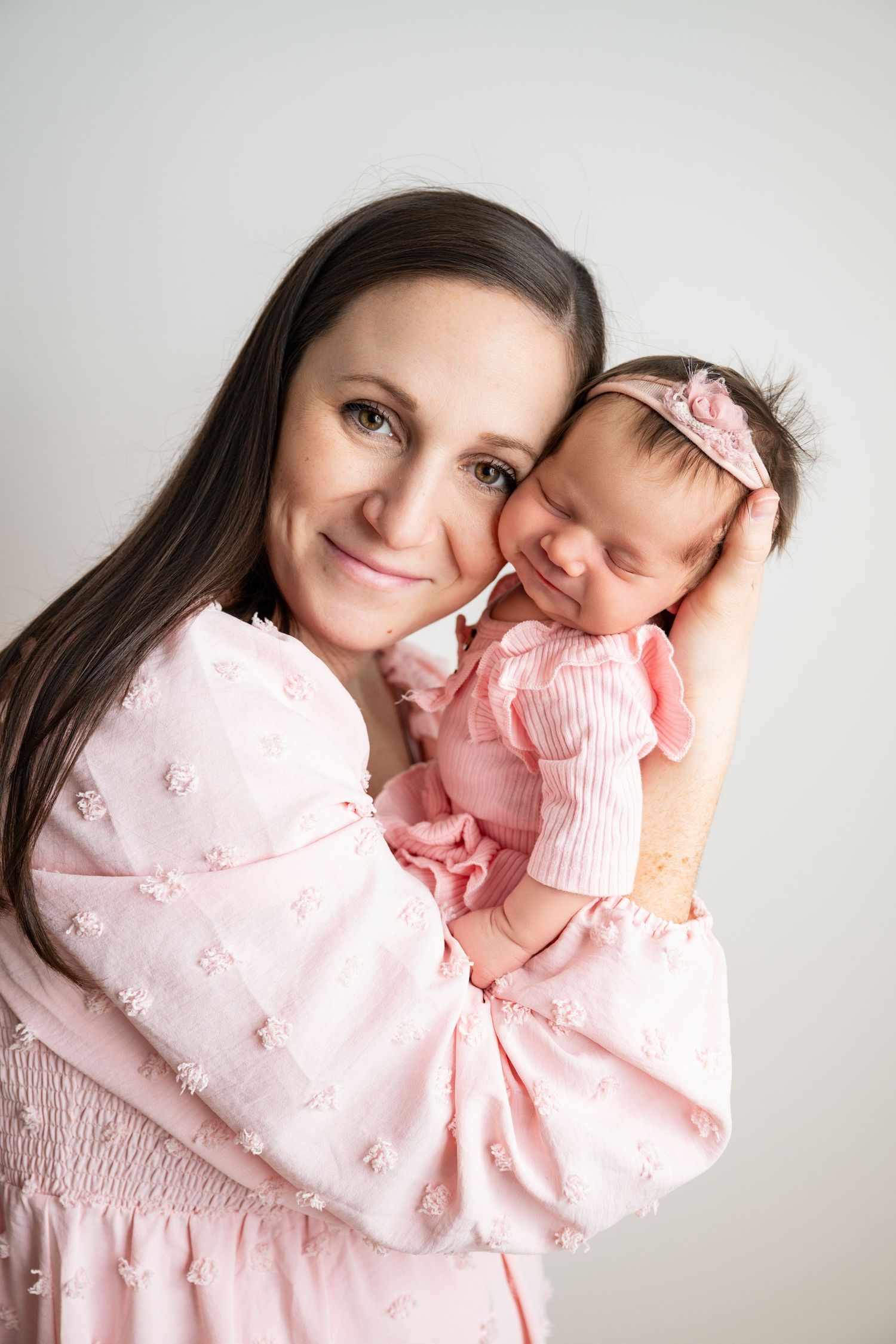 A smiling dark-haired woman in pink holds a sleeping newborn baby girl dressed in pink with a bow headband.