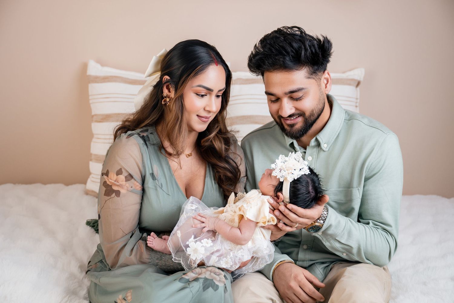 A couple in sage green outfits lovingly holds their newborn baby during a soft, neutral-toned newborn photography session.