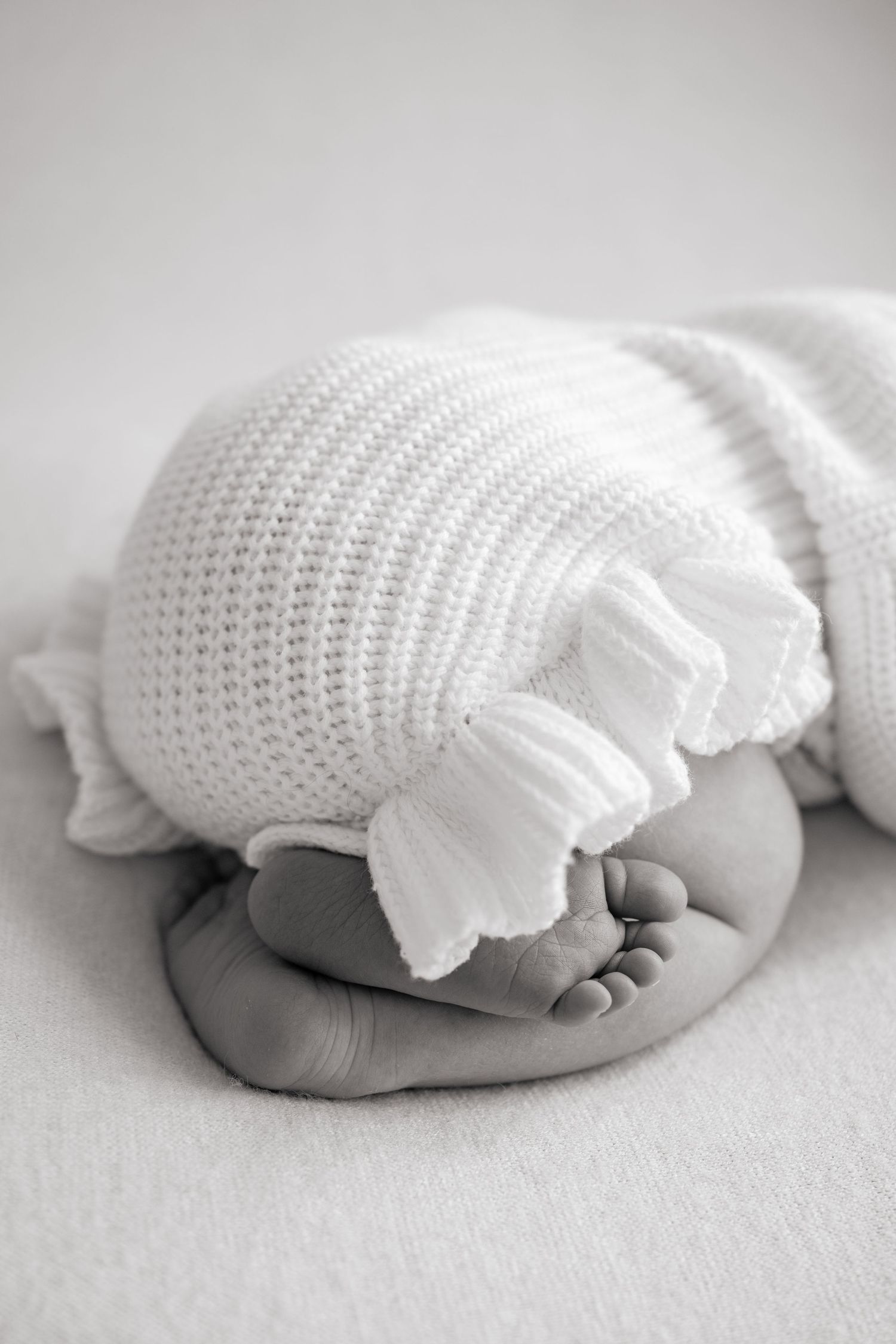 Close-up of a sleeping newborn baby's tiny feet and toes wrapped in a white knit blanket on a soft white background.