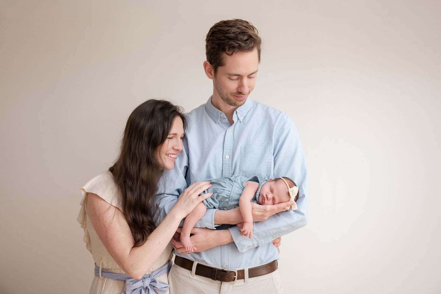 A young couple smiles lovingly at their newborn baby, with the father cradling the infant in a bright studio setting.