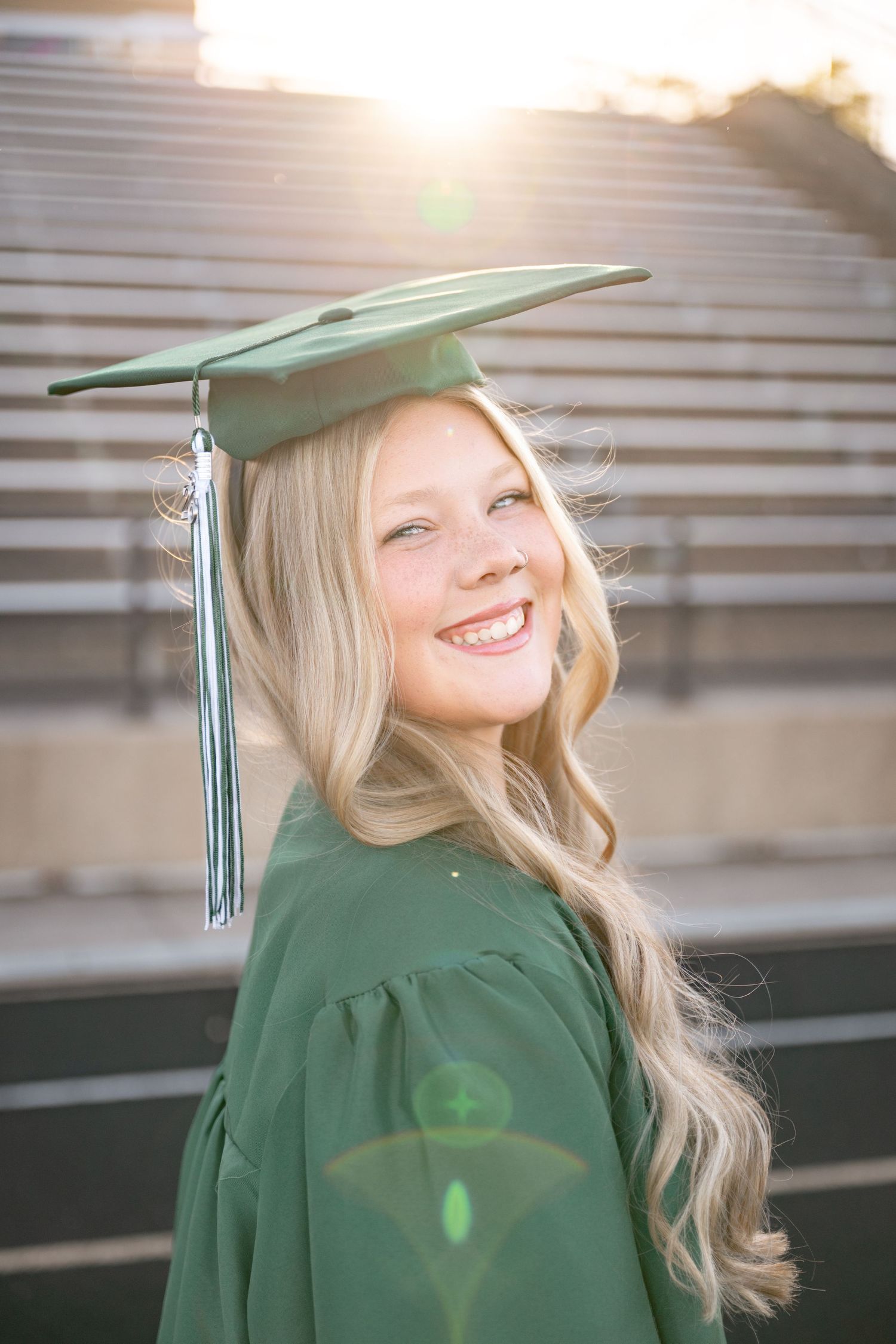 Smiling blonde graduate in green cap and gown poses outdoors in stadium with golden sunlight backdrop.