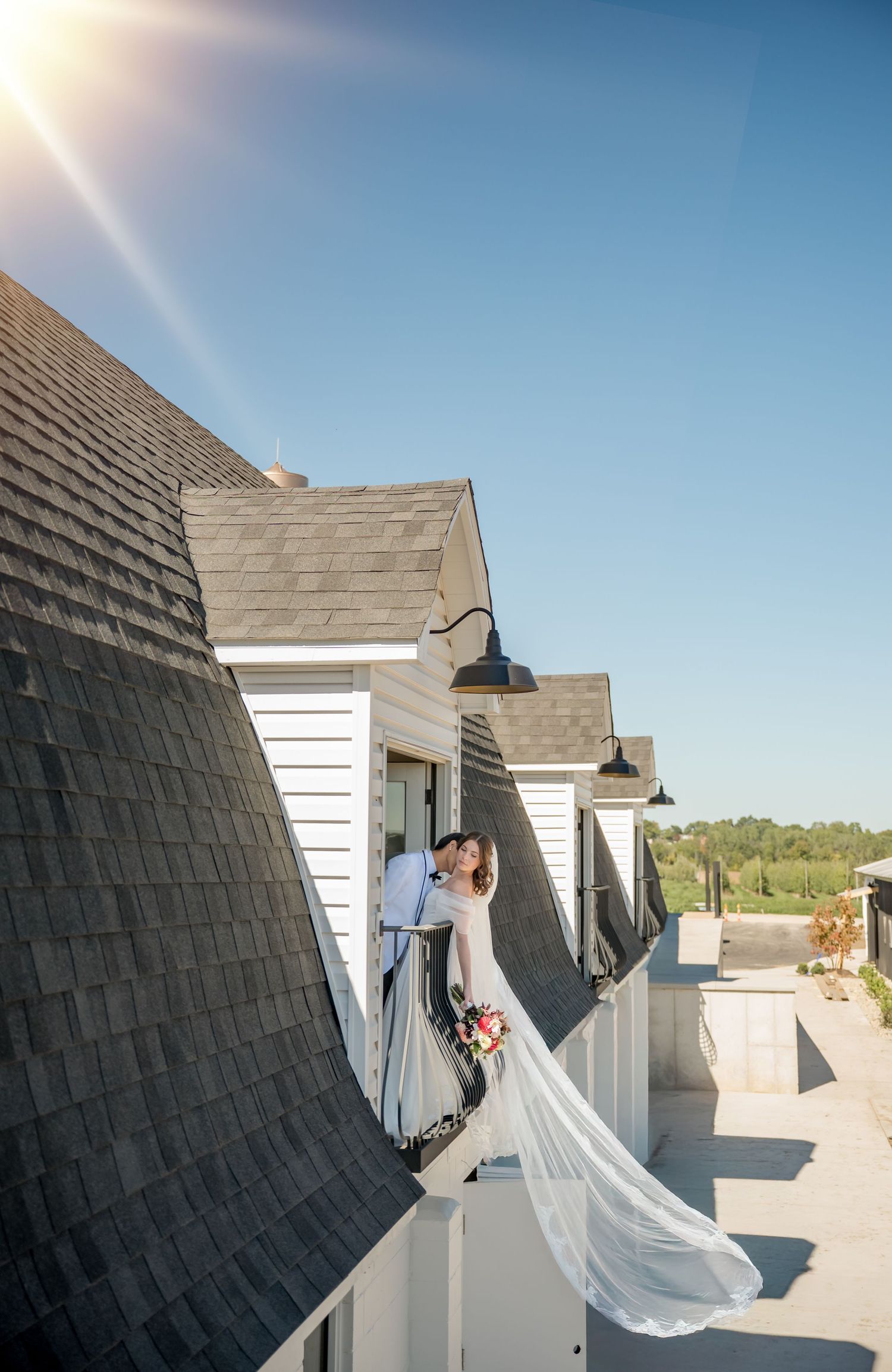 Bride and groom kissing on a sunlit rooftop balcony, wedding veil flowing in the breeze against a clear blue sky.