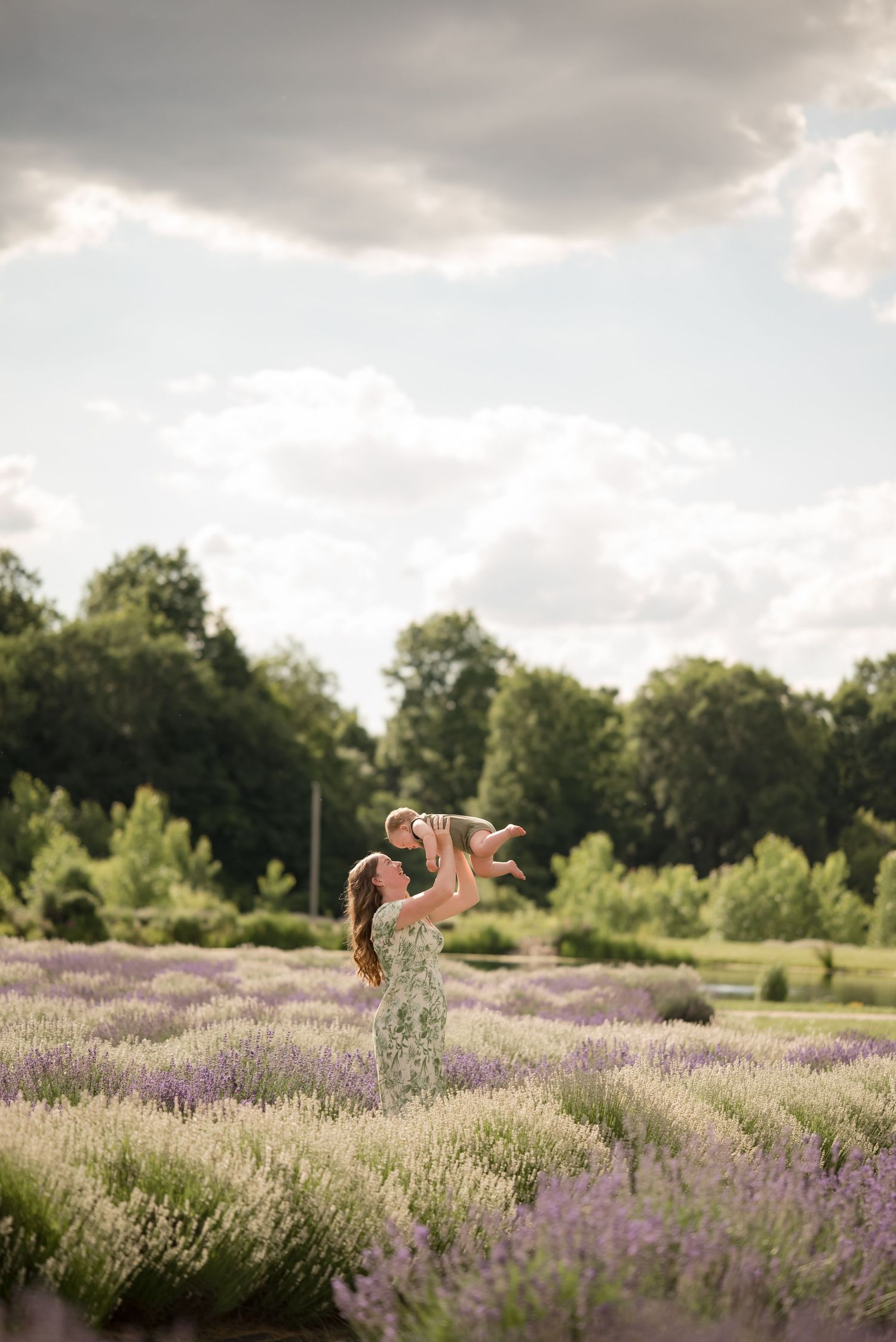 A joyful mother lifts her baby high in a lavender field, surrounded by lush trees under a cloudy sky.