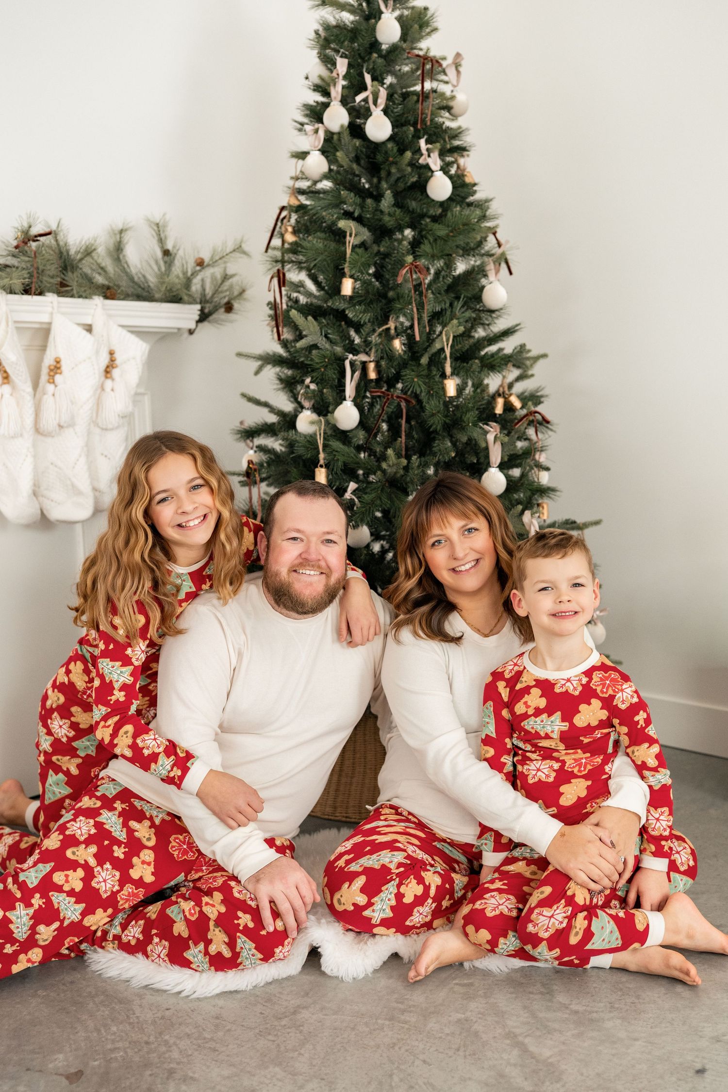 Family of four in matching red Christmas pajamas sitting together in front of decorated tree with stockings hung nearby.