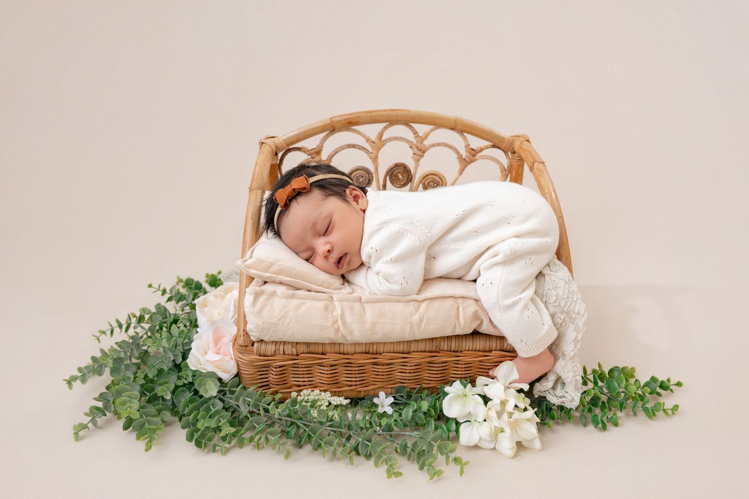 Sleeping newborn baby in white outfit on a wicker bed surrounded by green leaves and white flowers.
