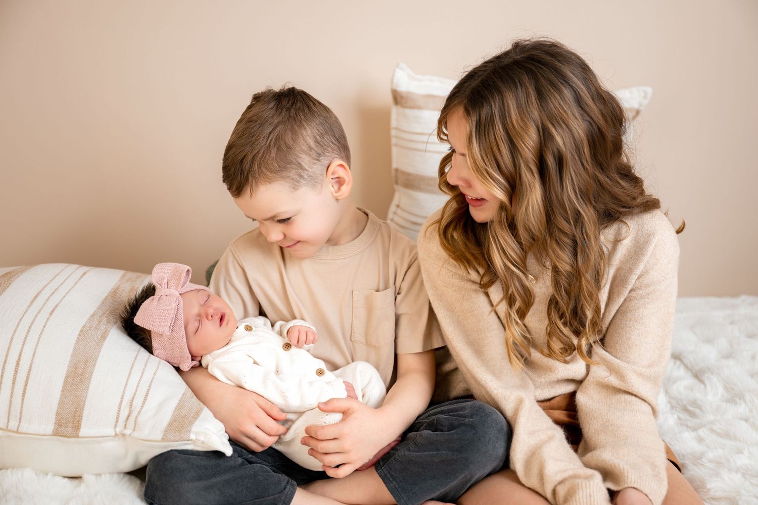 A mother and young boy sit together on a bed, lovingly gazing at a newborn baby wrapped in white.
