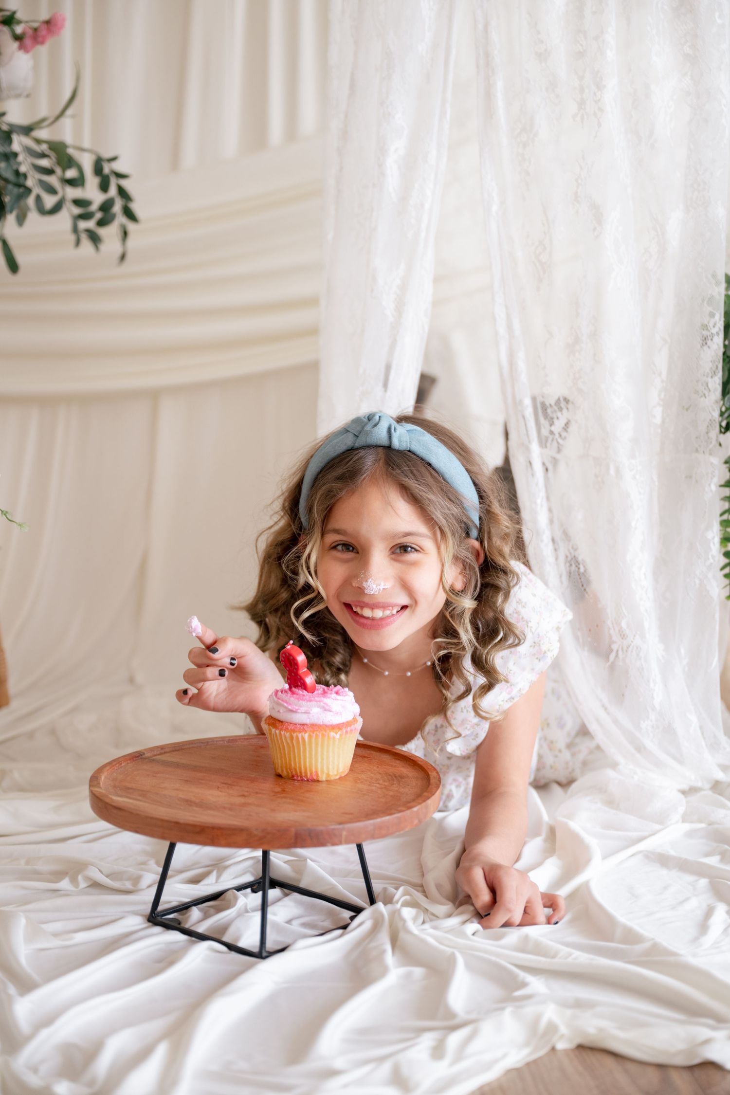 Smiling young girl in white dress with blue headband reaching for a pink frosted cupcake on a wooden cake stand.