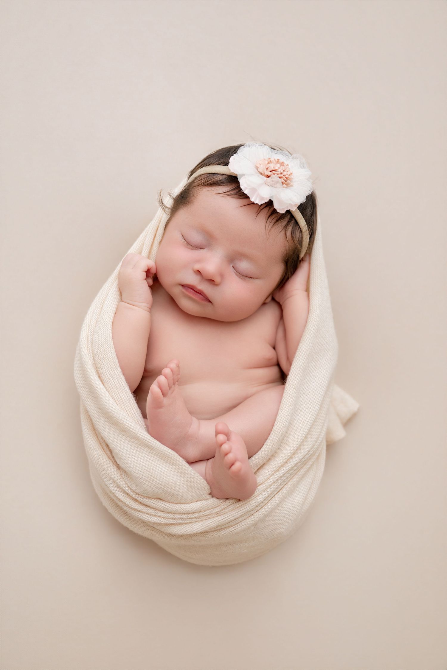 Sleeping newborn baby girl wrapped in cream swaddle with floral headband on beige background.