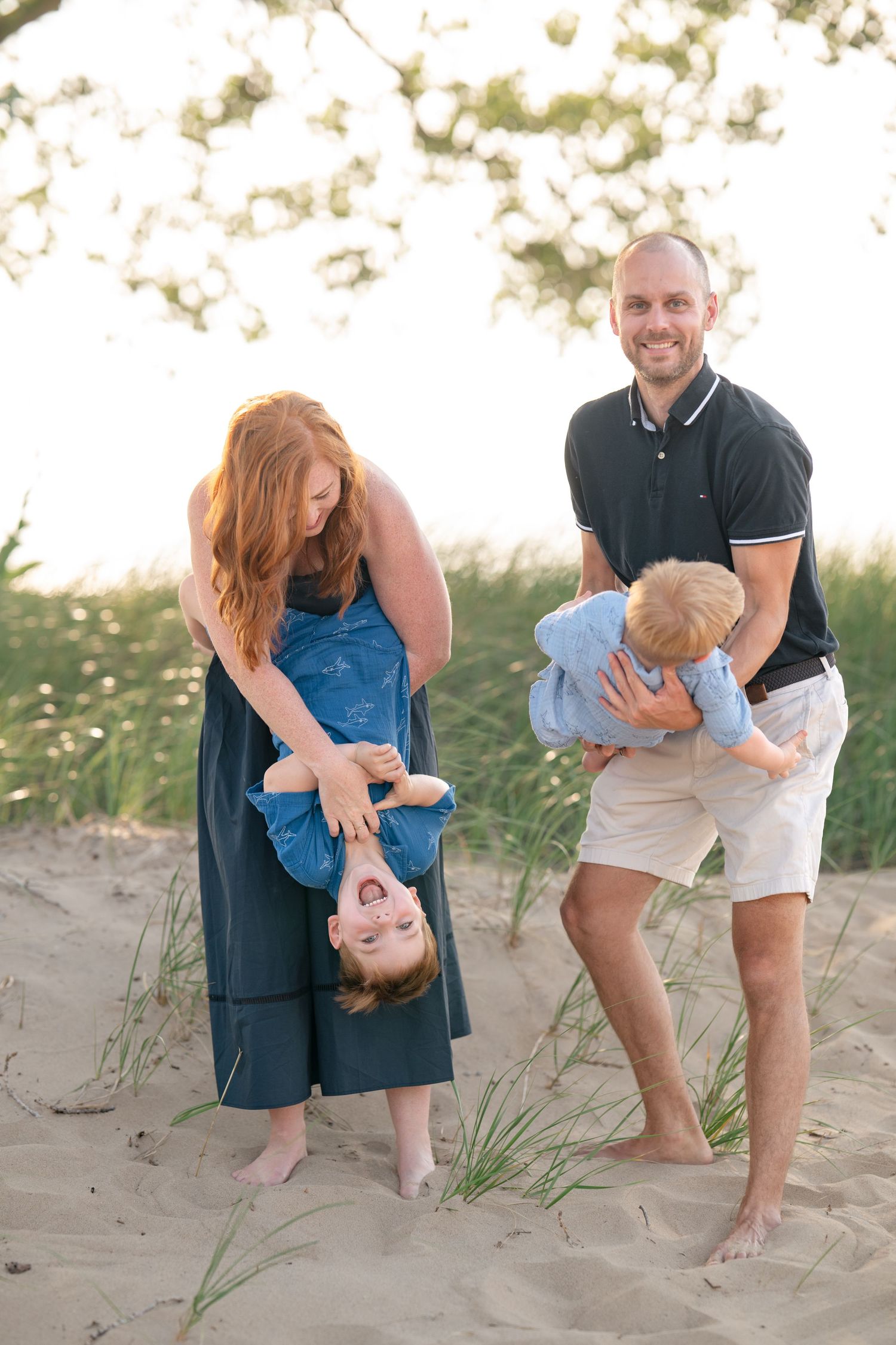Family of four playing on sandy beach dunes, mom holding toddler upside down while dad carries young child, golden hour light.