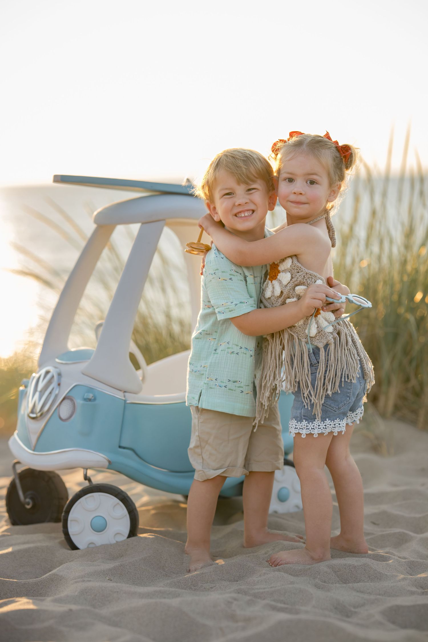 Two young children hugging and smiling on sandy beach beside a blue toy VW car at sunset.