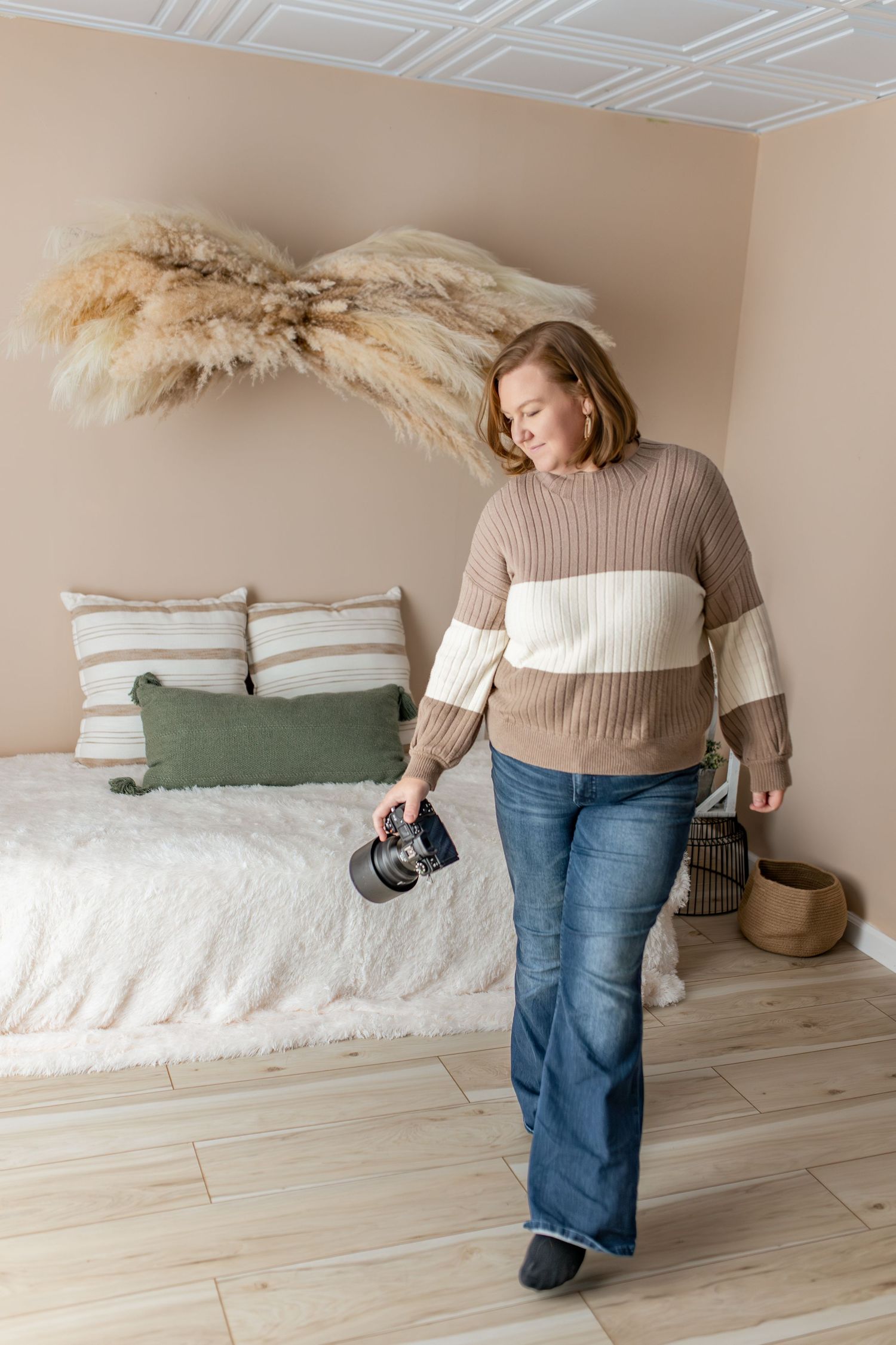 Woman in striped sweater and jeans holding a camera walks through a boho bedroom with pampas grass decor.