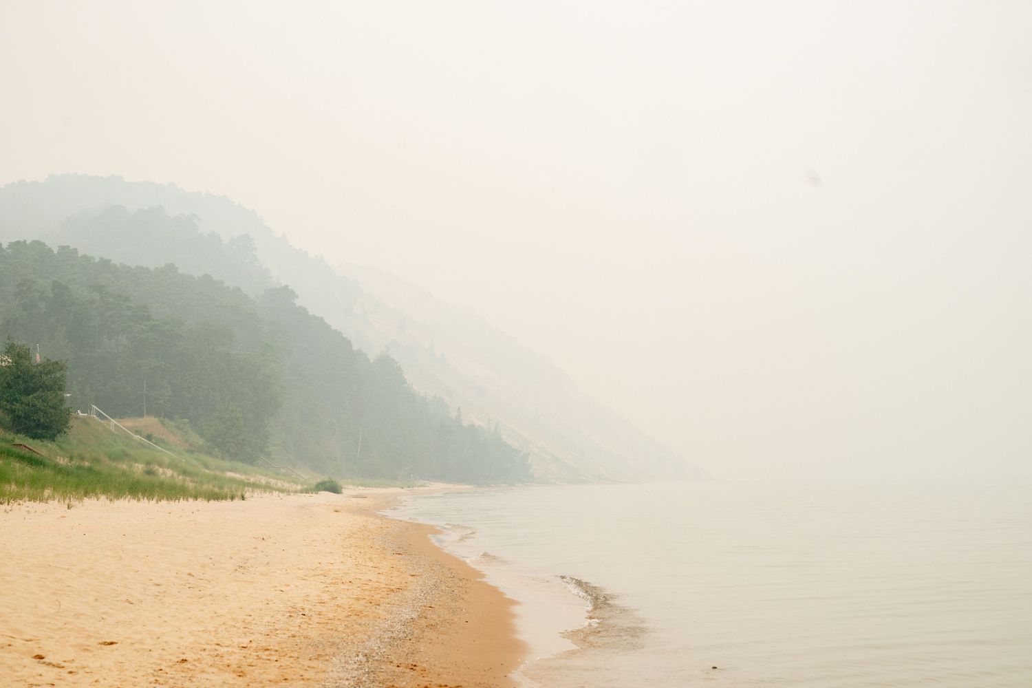 Misty sandy beach shoreline with lush green forested hills fading into hazy fog over calm water.
