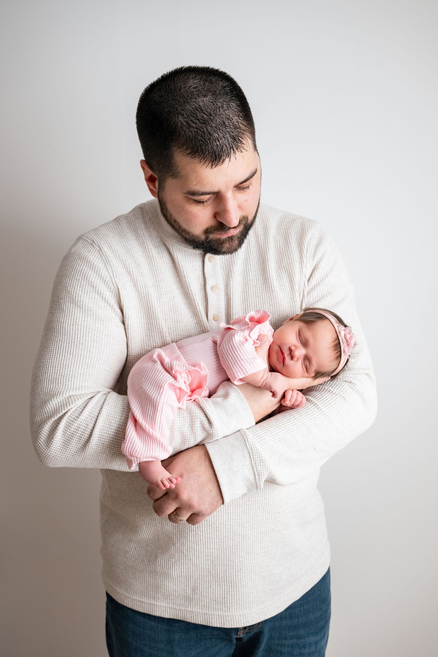 Father in beige sweater tenderly holds sleeping newborn baby girl dressed in pink outfit with bow headband.