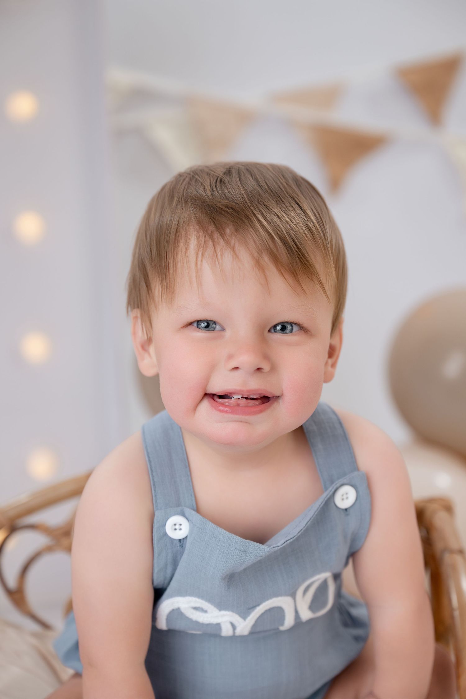 An adorable smiling toddler boy in a blue romper sits in front of festive birthday balloons and string lights.