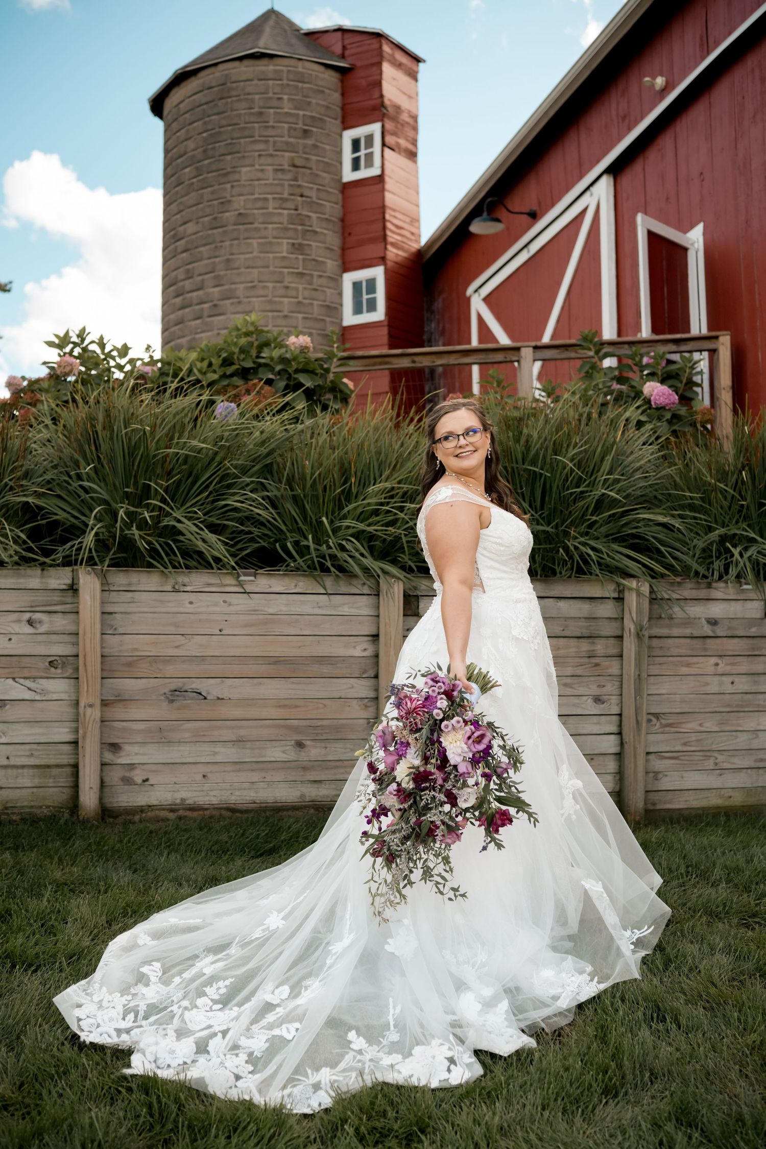 Bride in white lace gown holding purple bouquet poses before red barn and silo on sunny wedding day.