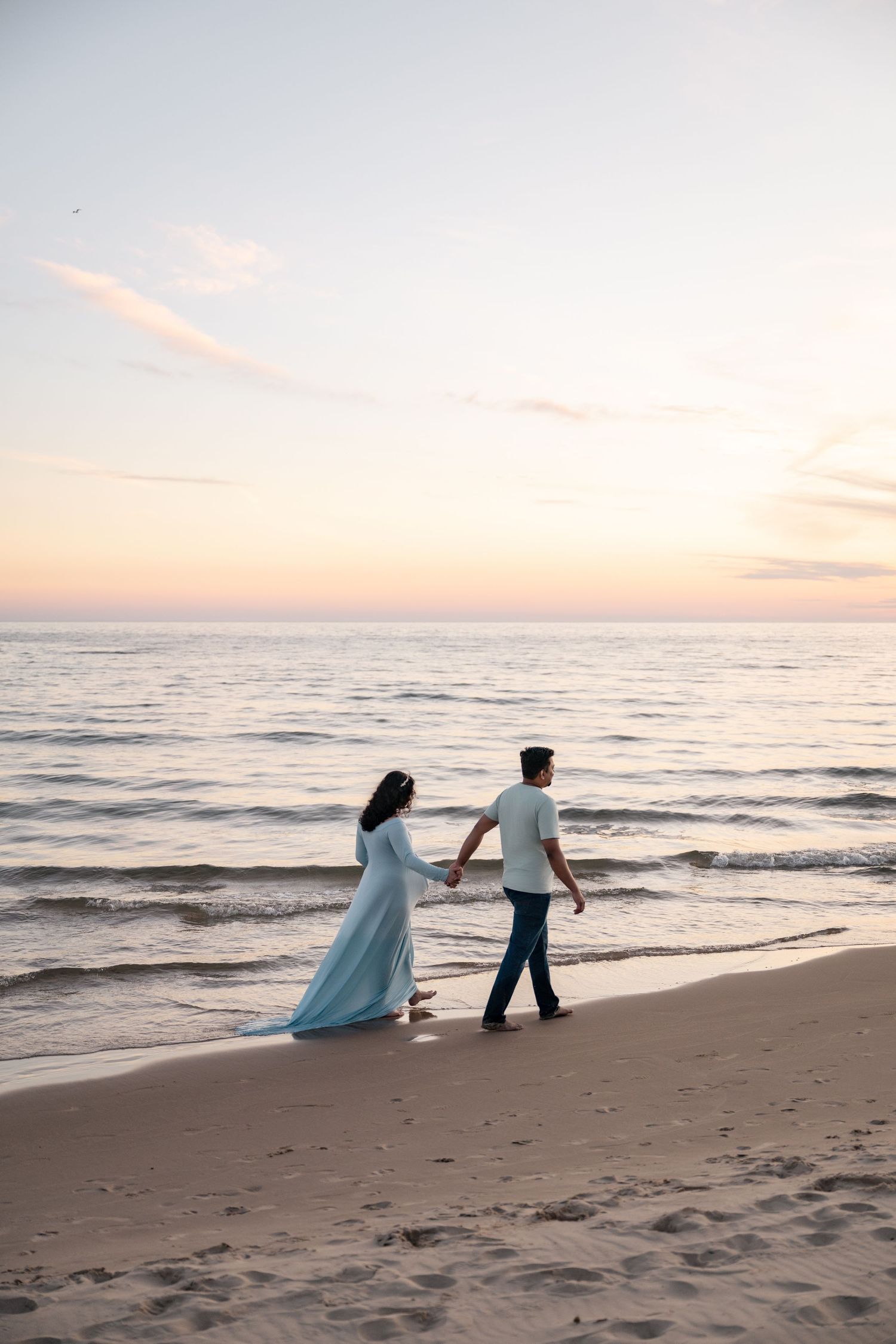 Couple holding hands walking on sandy beach at sunset, woman in flowing light blue dress, romantic seaside stroll.