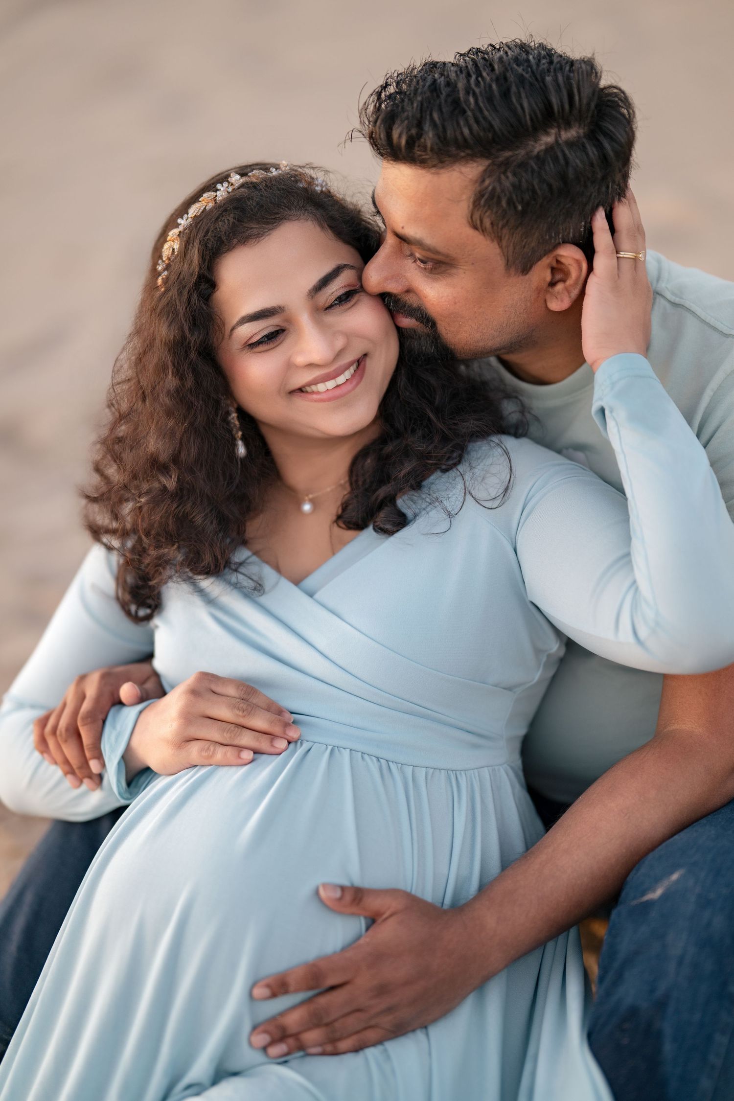 A smiling pregnant woman in a light blue dress is kissed by her husband while cradling her baby bump outdoors.