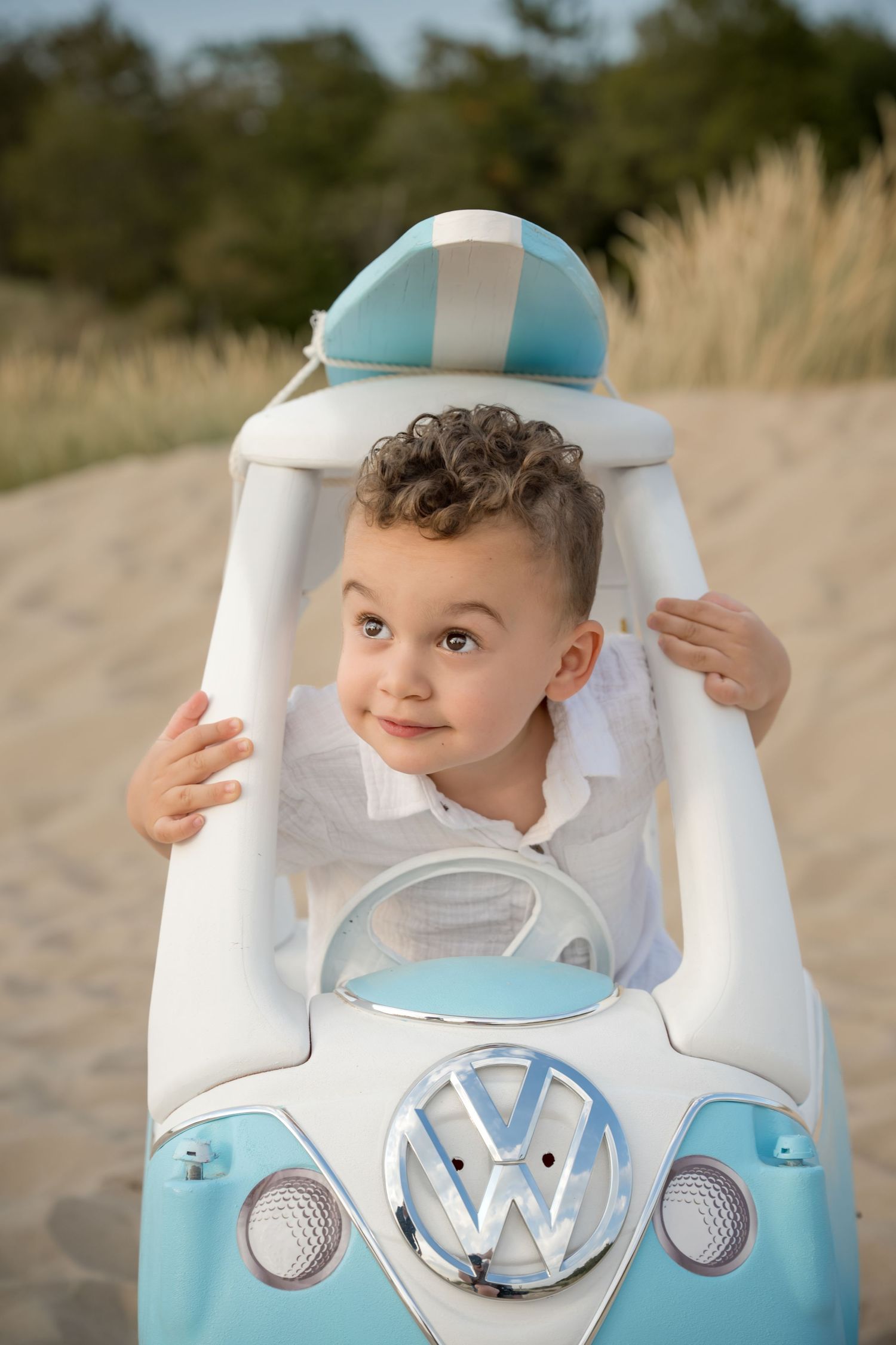 Young curly-haired boy smiling inside a blue and white toy Volkswagen van on a sandy beach.